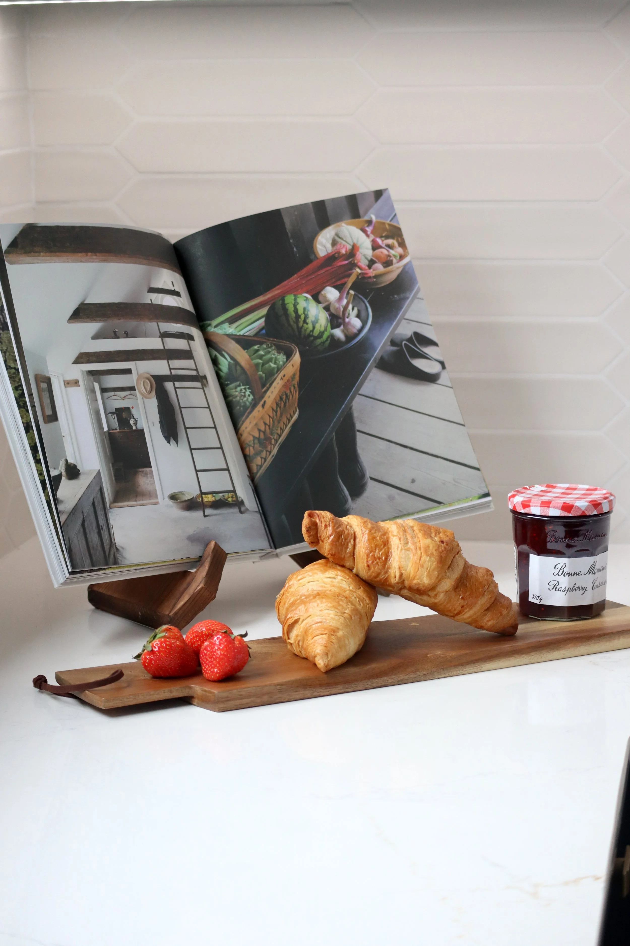 A wooden cutting board with three strawberries, two croissants, and a jar of raspberry jam. Behind the board, an open cookbook displays images of vegetables and a rustic kitchen. The background features white tiled wall.