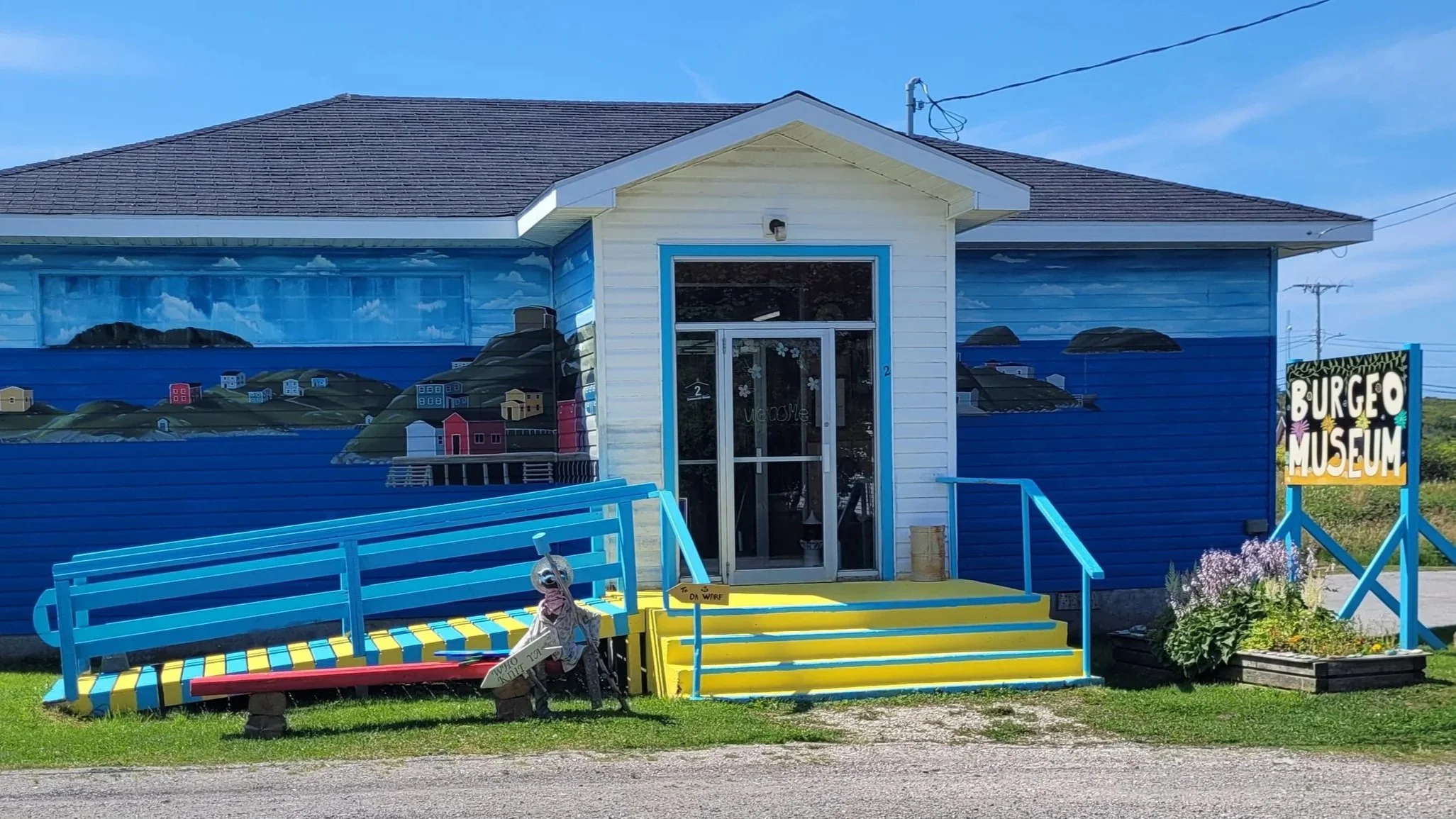 The exterior of the Burgeo Museum, a small white building with blue accents, featuring a colorful mural of a coastal scene with houses and hills on the side. There is a blue and yellow ramp and stairs leading to the entrance, decorated with a sign and flowers nearby.