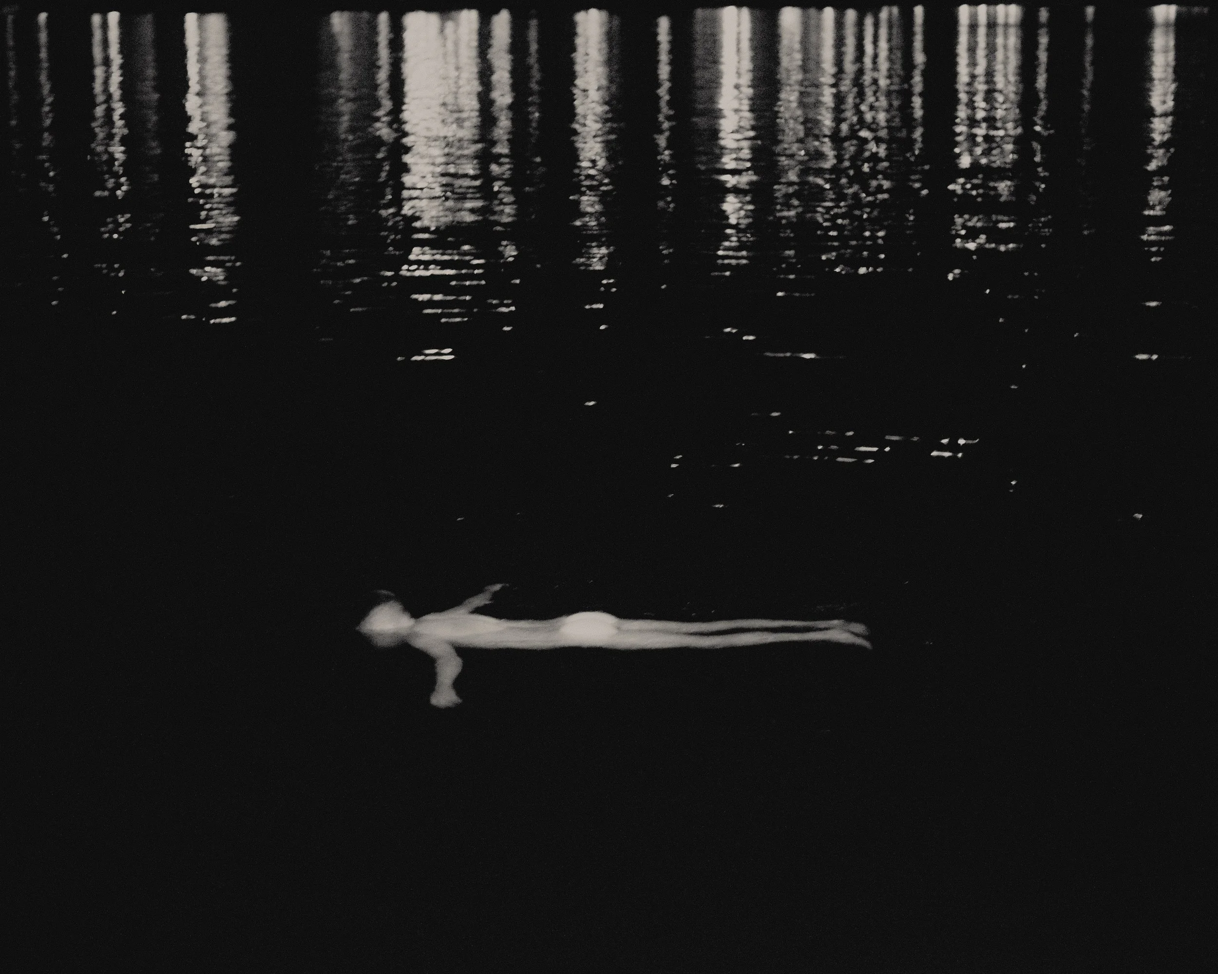 Una figura humana flotando en un cuerpo de agua oscuro, con reflejos de luces en la superficie del agua.
