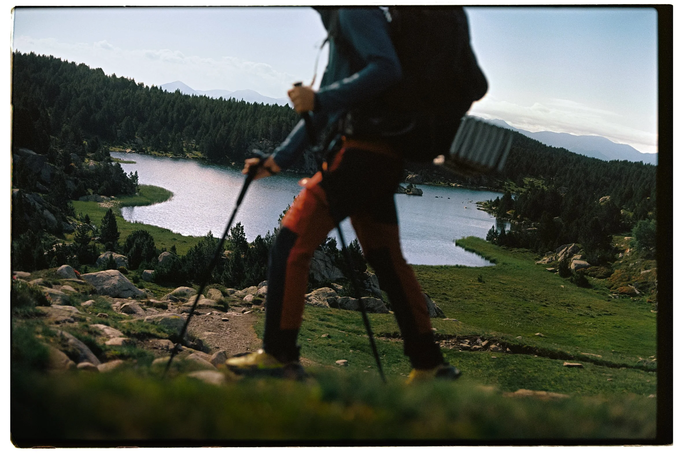 Persona haciendo senderismo en una ruta de montaña con vista a un lago, rodeada de árboles y montañas.