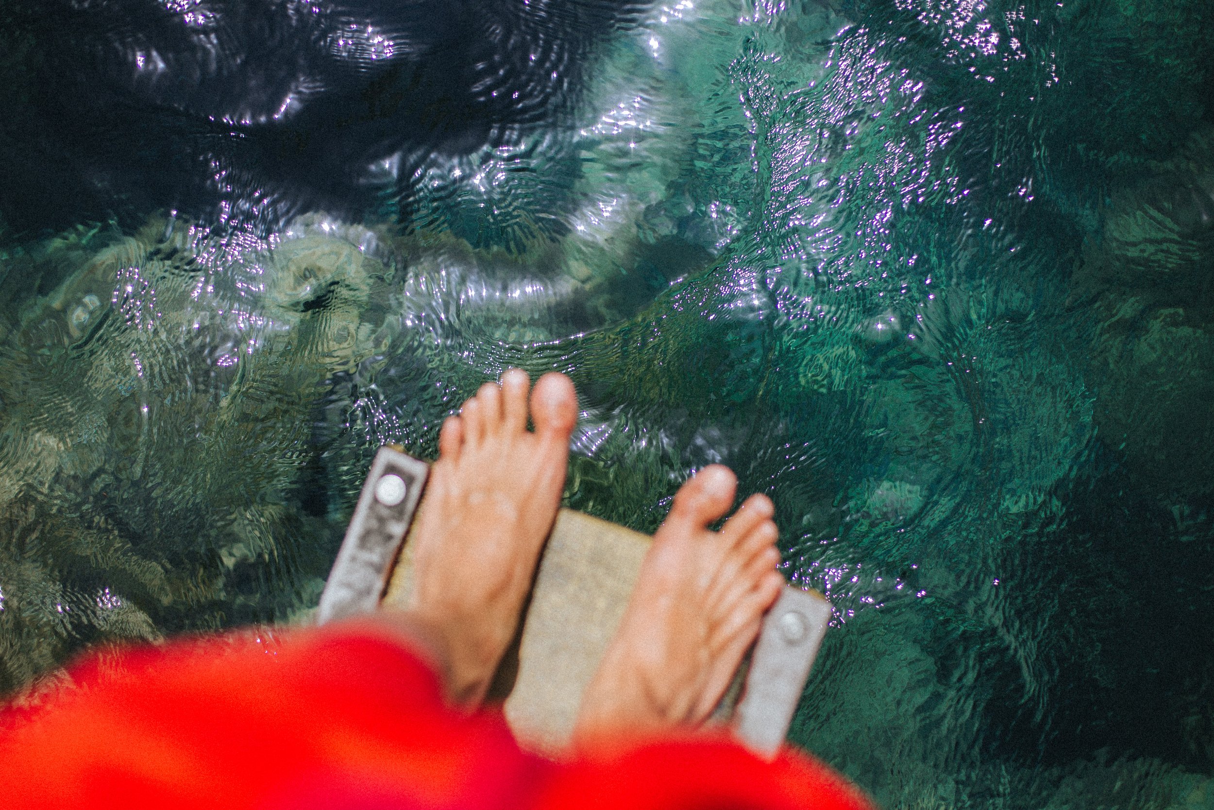 Pies de una persona en una tabla en el borde de un cuerpo de agua, probablemente un río o lago, con agua clara jade y reflejos de luz.