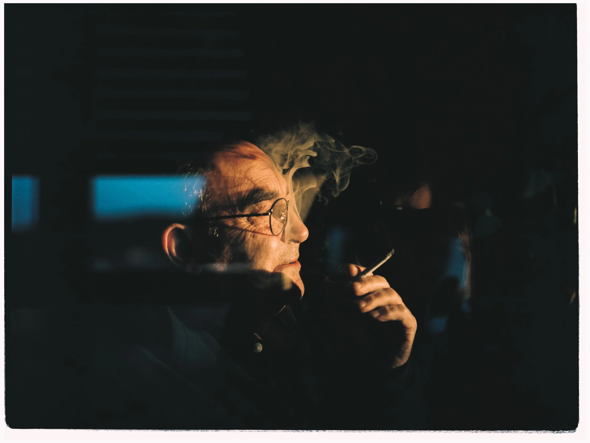 Hombre con gafas fumando un cigarrillo en un ambiente oscuro con humo en el aire.