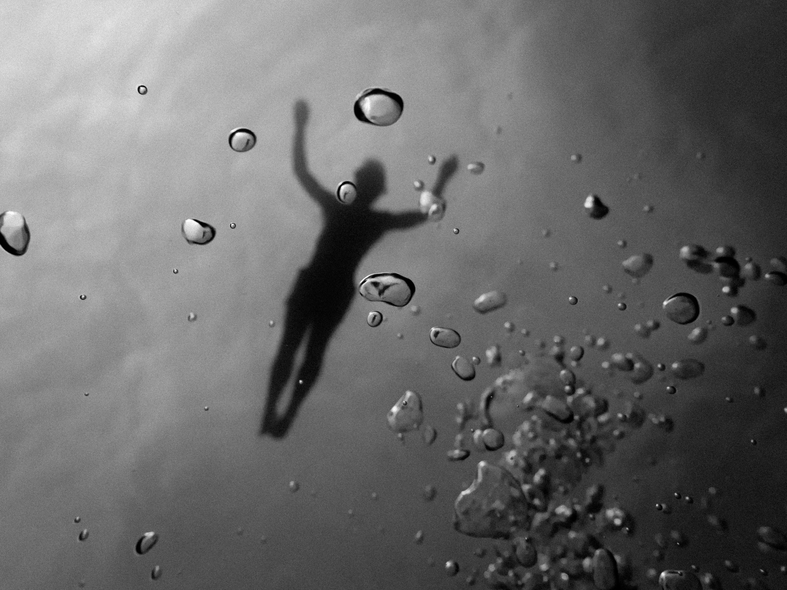 Silueta de una persona joven suspendida en agua, vista desde abajo, rodeada de burbujas. La imagen es en blanco y negro.