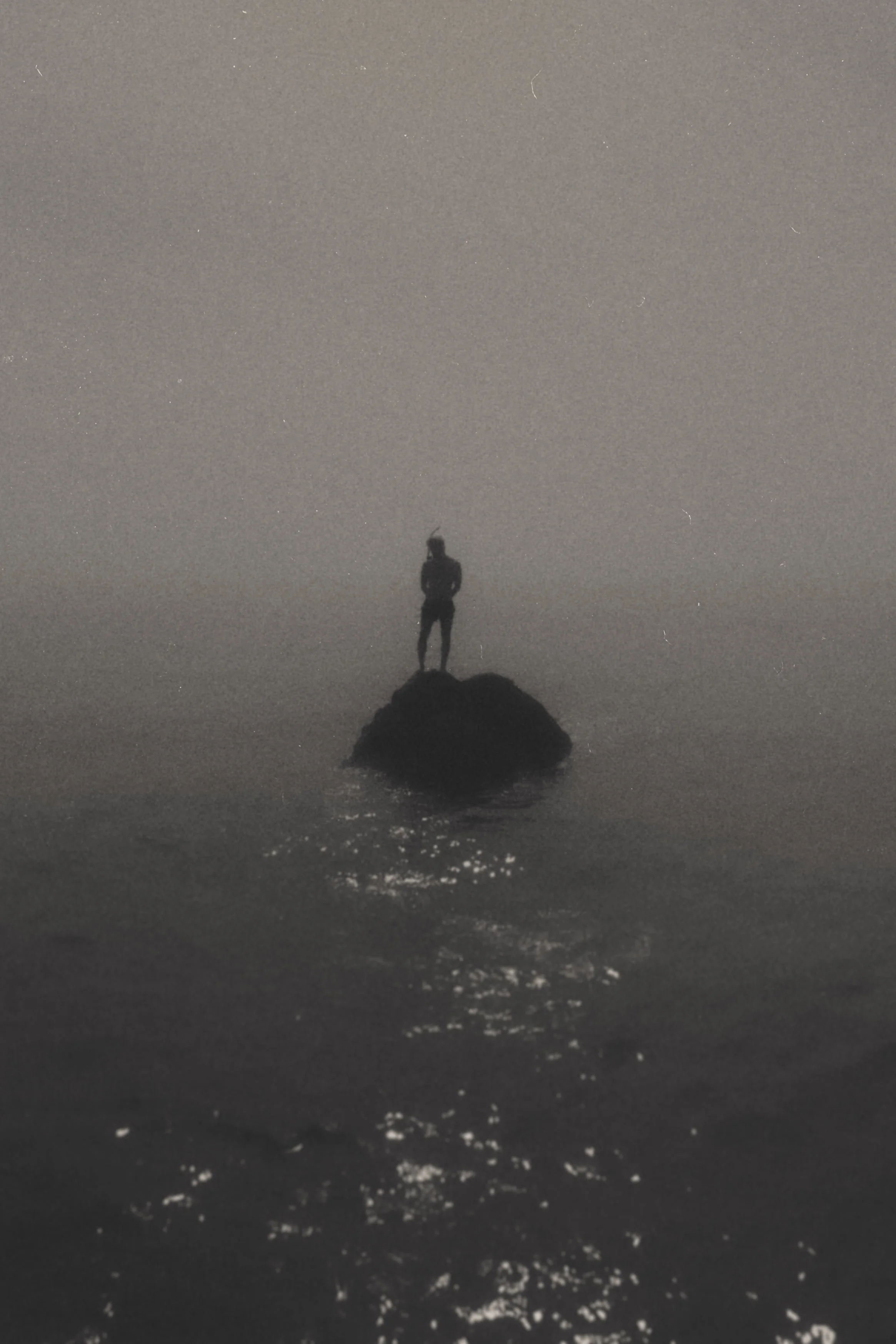 Una persona en pie sobre una roca en el mar durante la noche, con un cielo oscuro y sin estrellas.