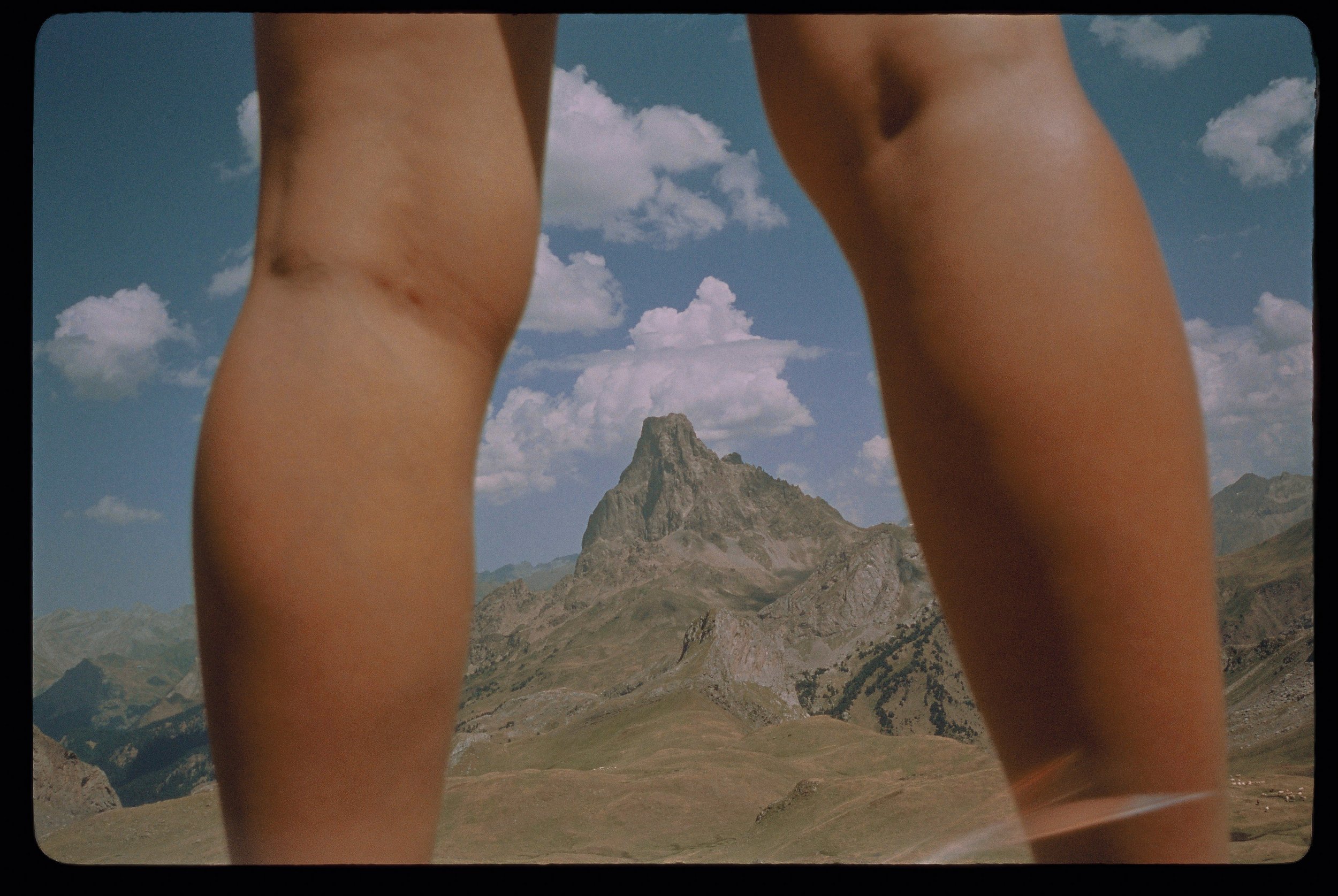 Dos piernas de una persona en primer plano, con un paisaje montañoso y una cumbre rocosas en el fondo, bajo un cielo con nubes.
