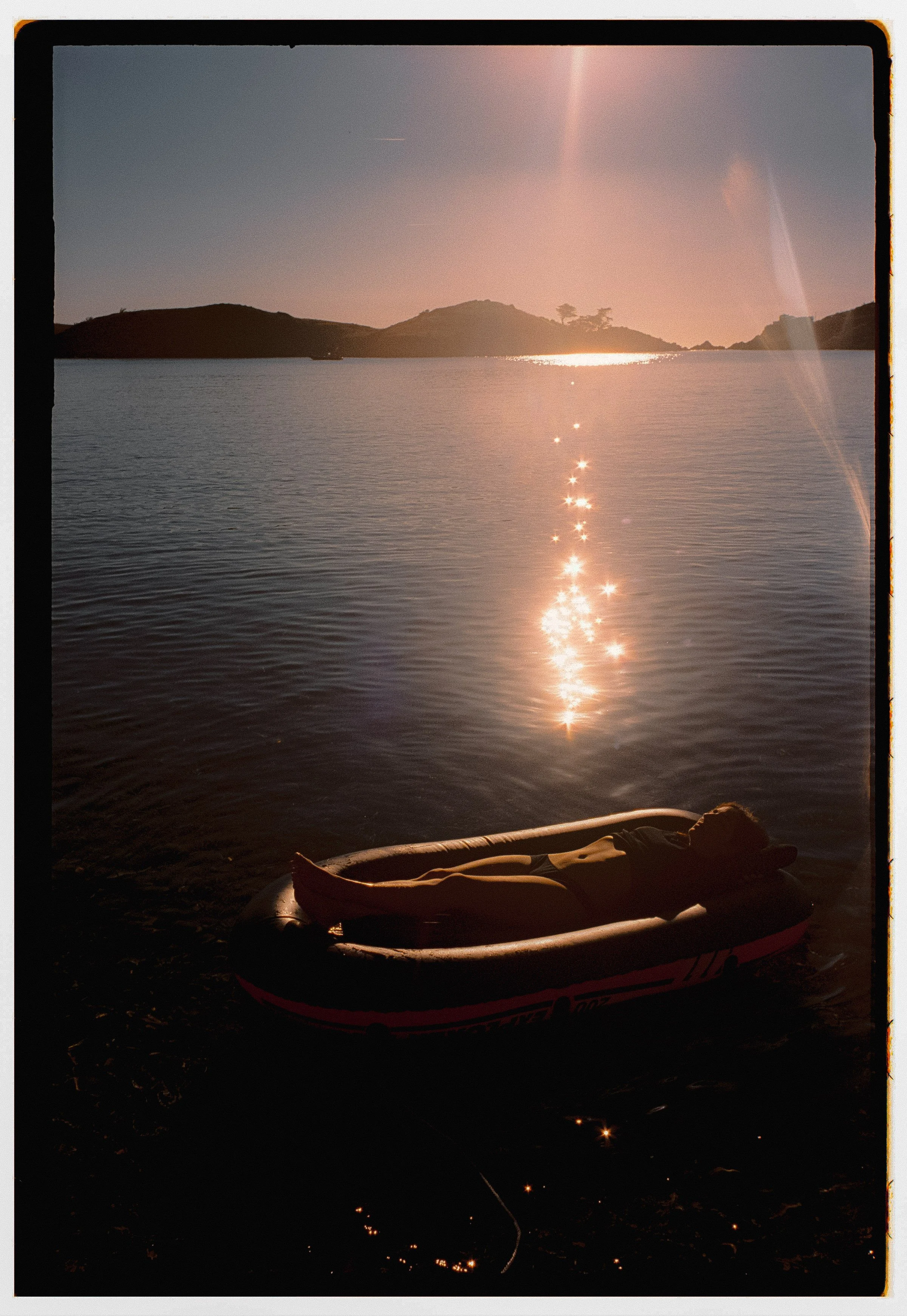 Persona durmiendo en una cama inflable junto a un lago al atardecer, reflejando el sol en el agua y un paisaje de colinas y árboles en el fondo.