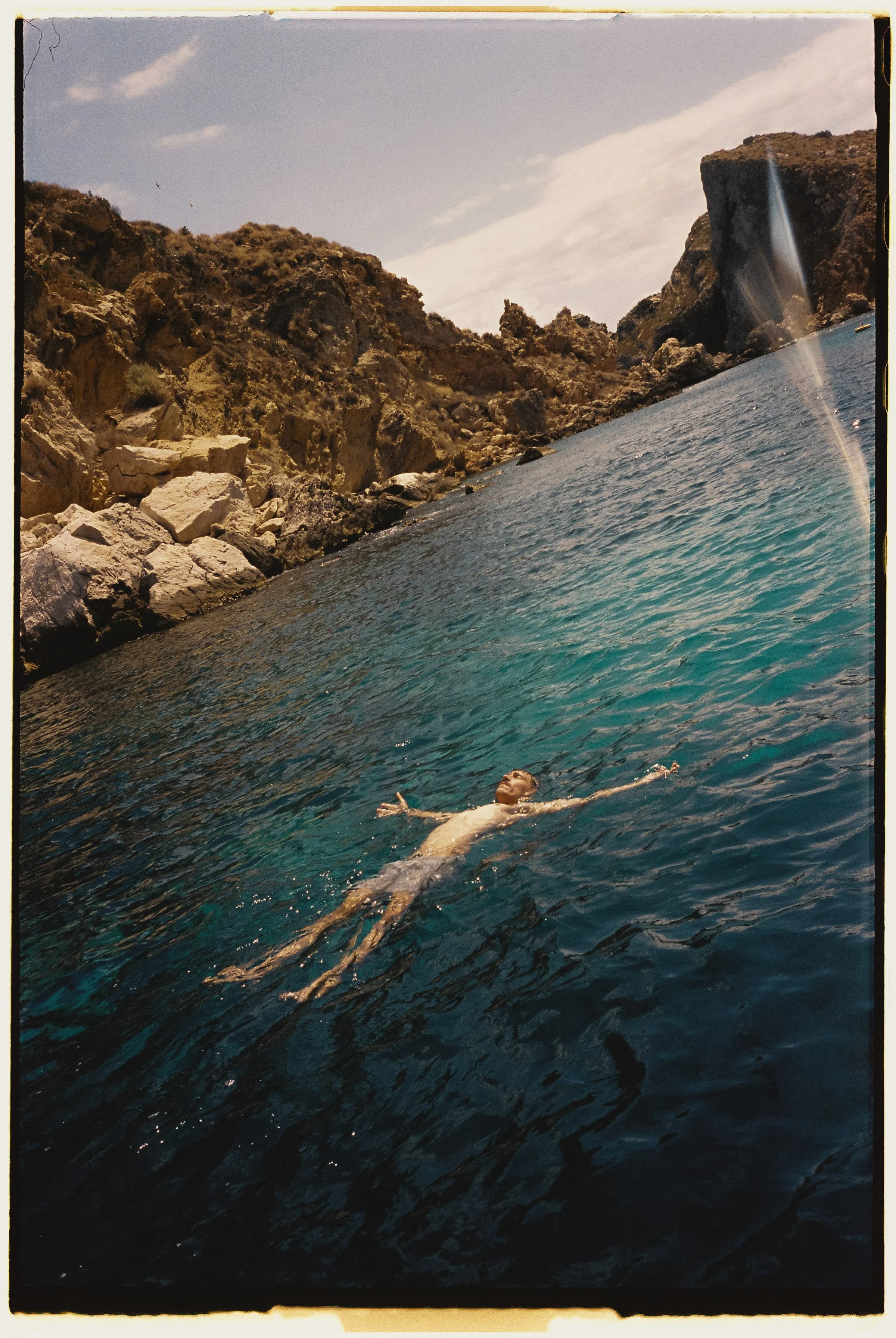 Persona nadando en el agua junto a rocas y un acantilado con una cascada en la distancia.