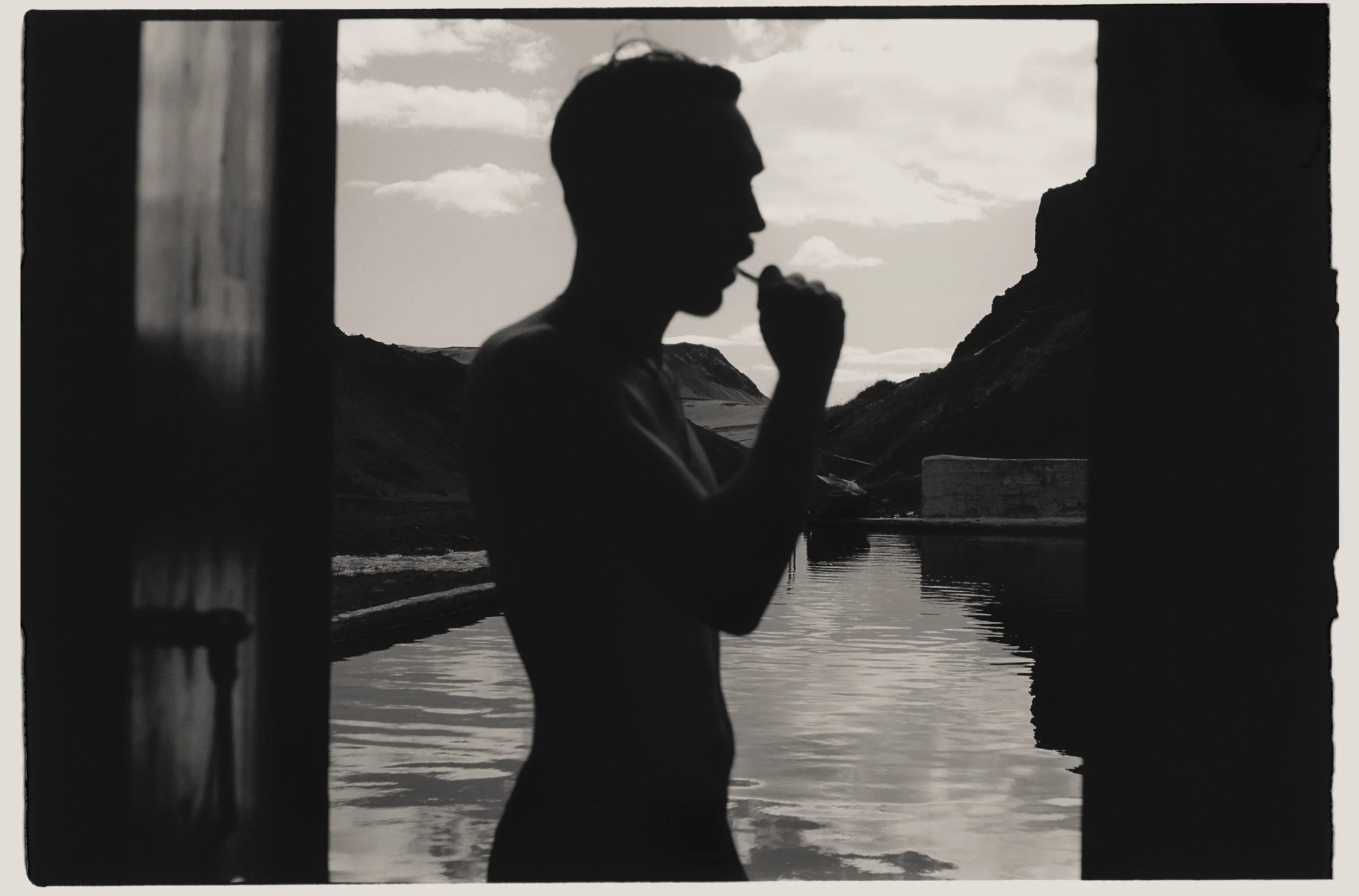 Silhouette de un hombre joven tomando una masticando con una pajilla, visto desde una ventana con fondo de paisaje montañoso y lago.
