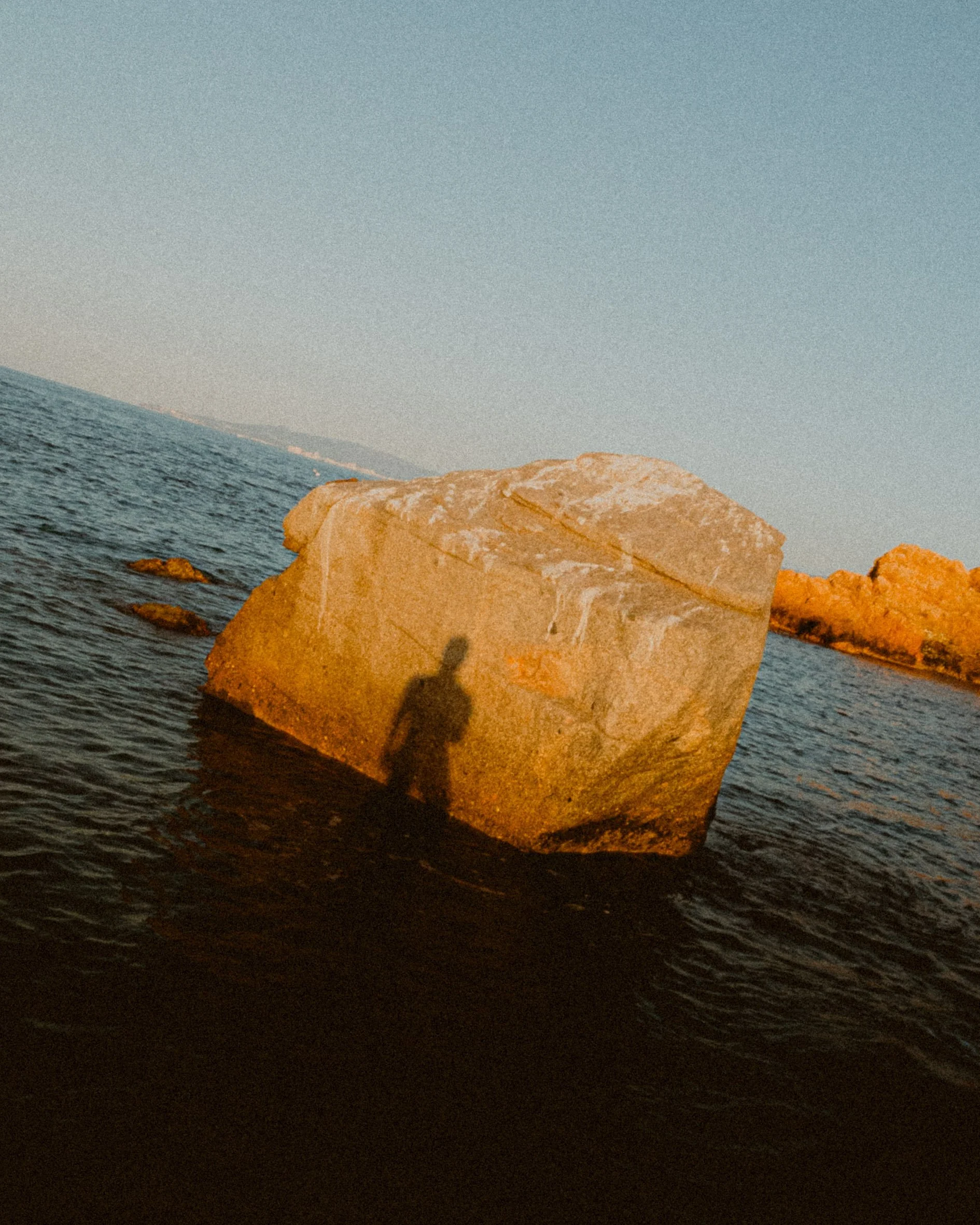 Una roca grande en el agua con la sombra de una persona proyectada sobre ella, al atardecer.