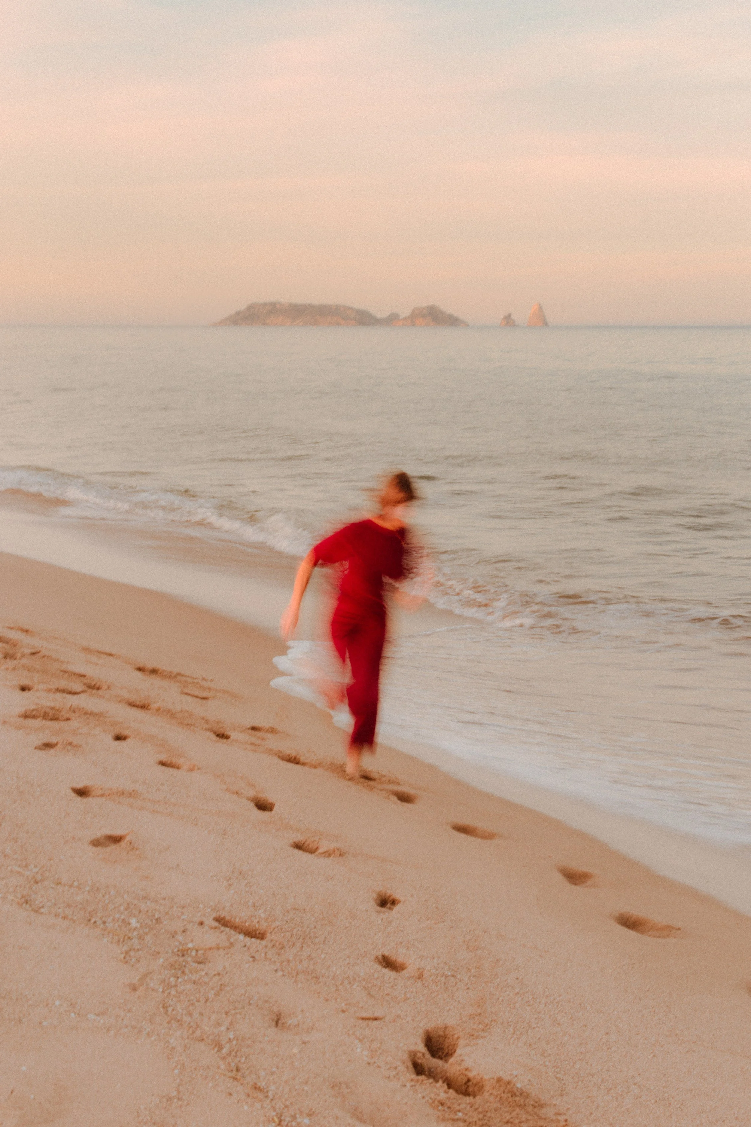 Una persona con ropa roja caminando descalza por la orilla de la playa con el mar al fondo y un par de islas en la distancia.
