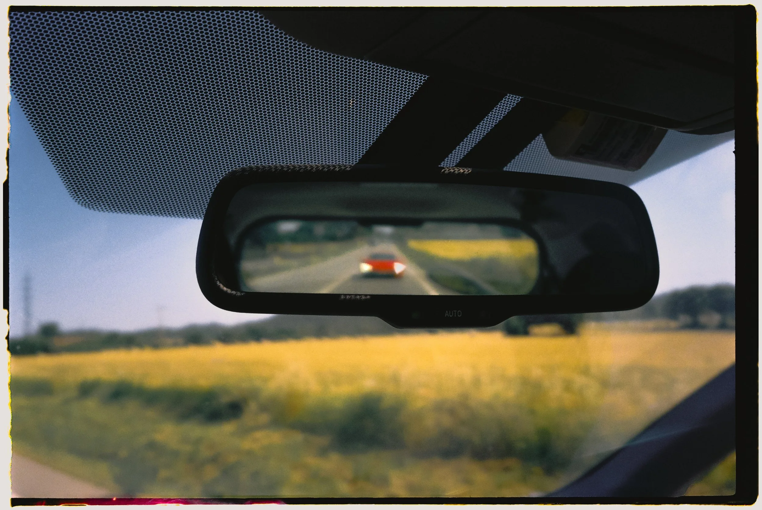 Vista desde el interior de un automotor, mostrando un espejo retrovisor con un camino y un vehículo gris detrás, campos verdes y cielo claro al fondo.