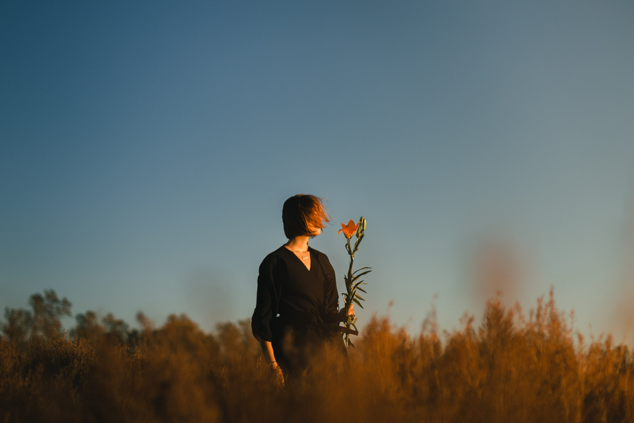 Una mujer con cabello corto y vestido negro sostiene un ramo de flores en un campo durante el atardecer.