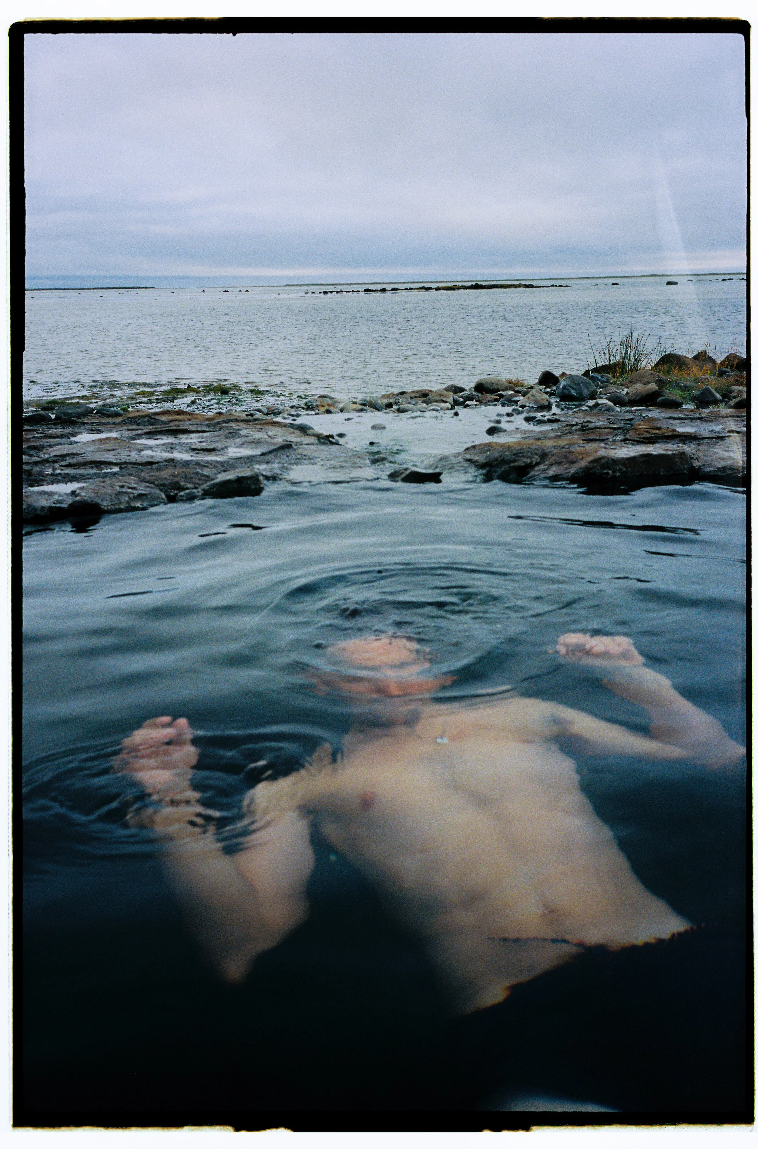 Persona desnuda flotando en el agua de un río o lago, con paisajes de piedras y agua en el fondo y un cielo nublado.