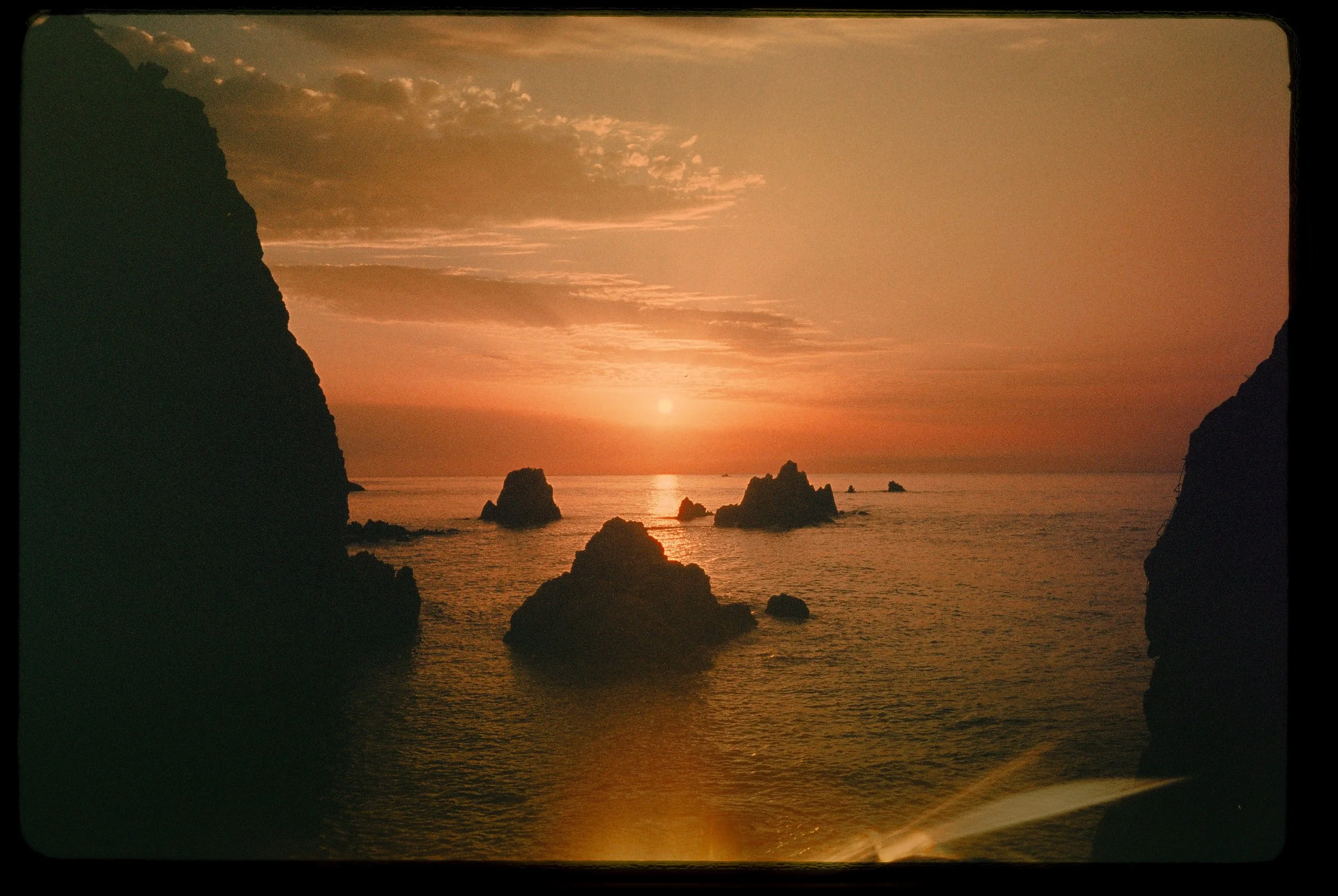 Atardecer sobre el océano con rocas en el agua, cielo con nubes y sol en el horizonte.