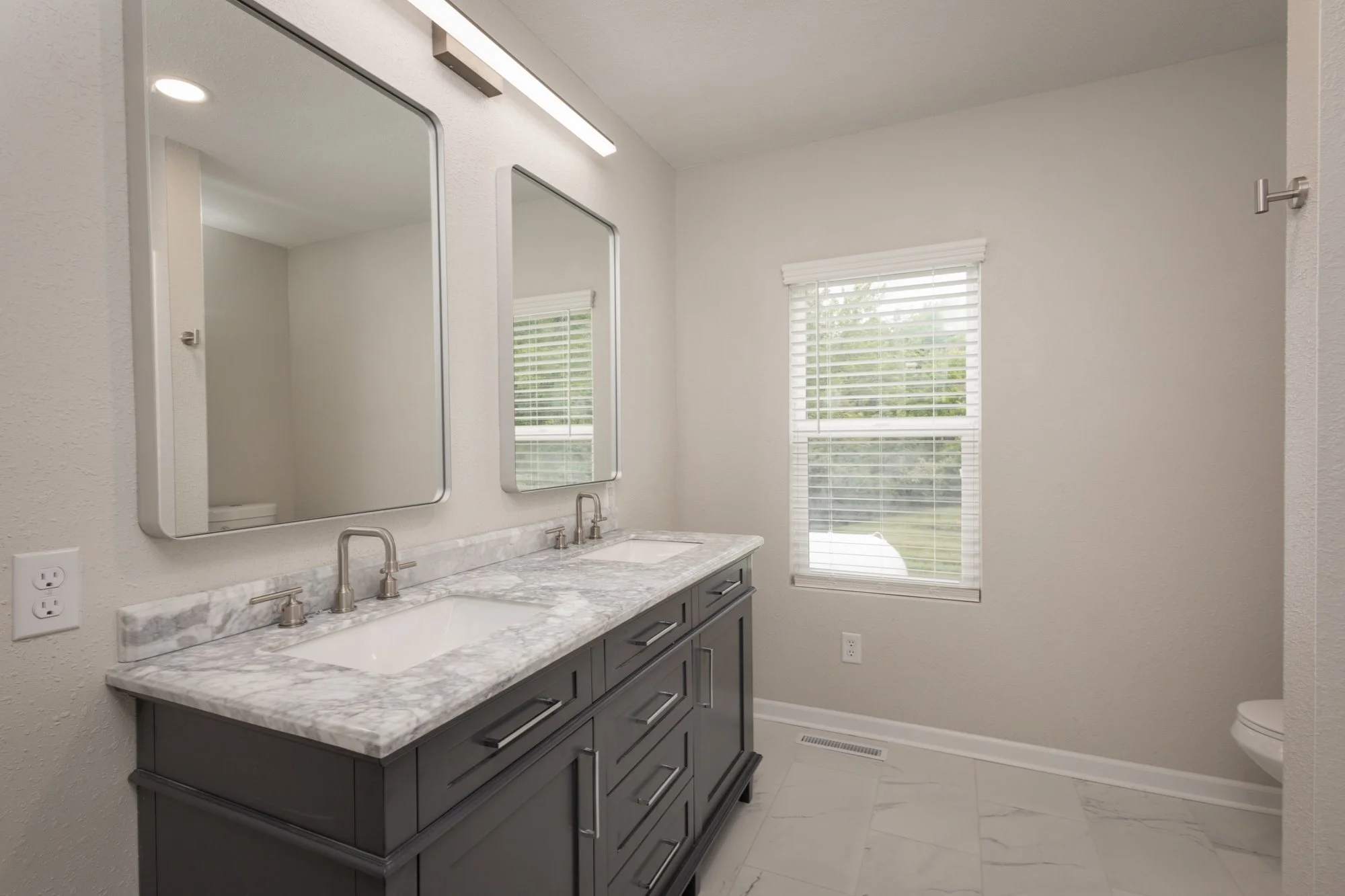 Bathroom with a double marble-sink vanity, two mirrors, and a window with blinds, featuring a gray cabinet and white walls.