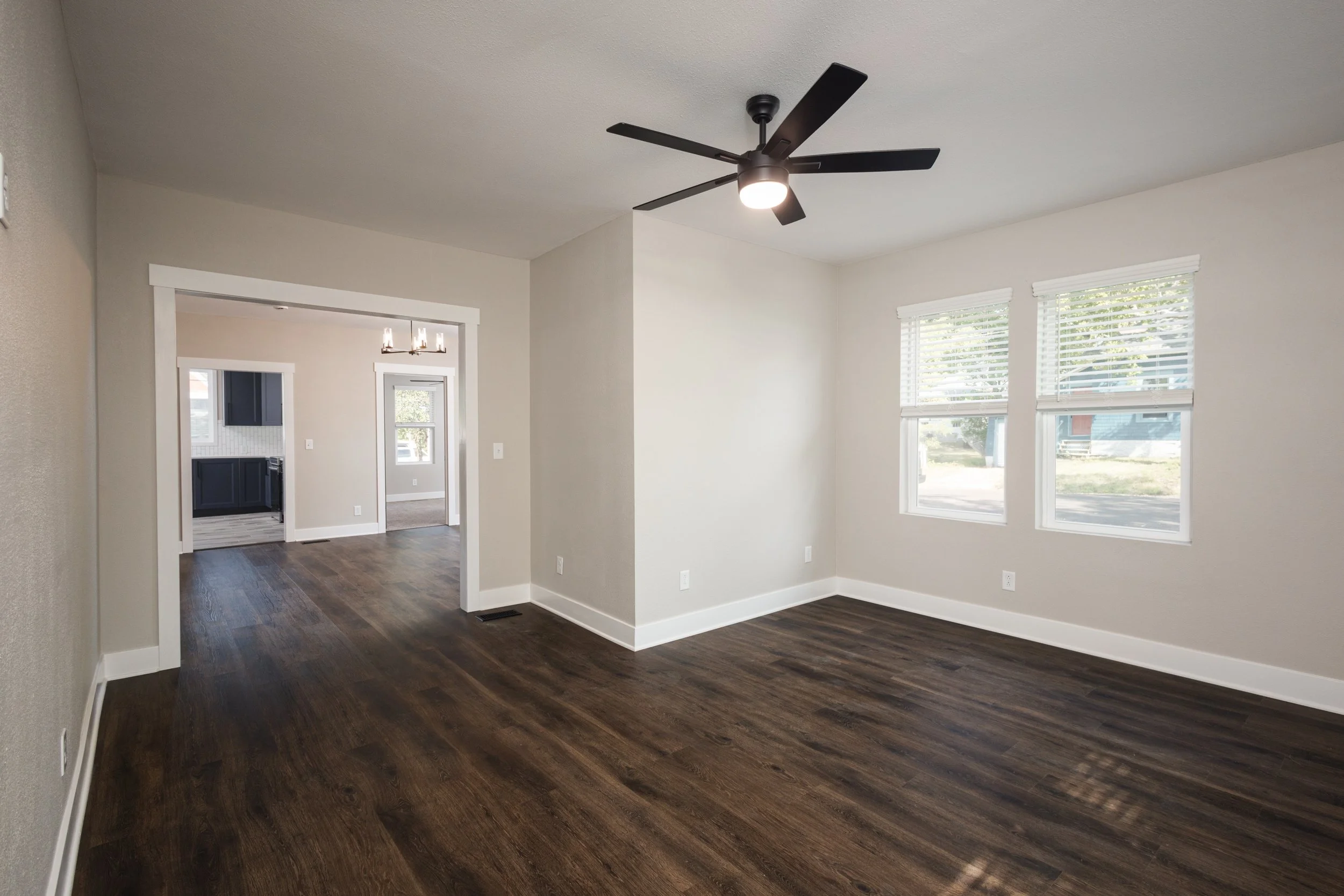 Empty living room with dark wood floors, white walls, two windows with blinds, a ceiling fan, and a view into a kitchen and dining area.