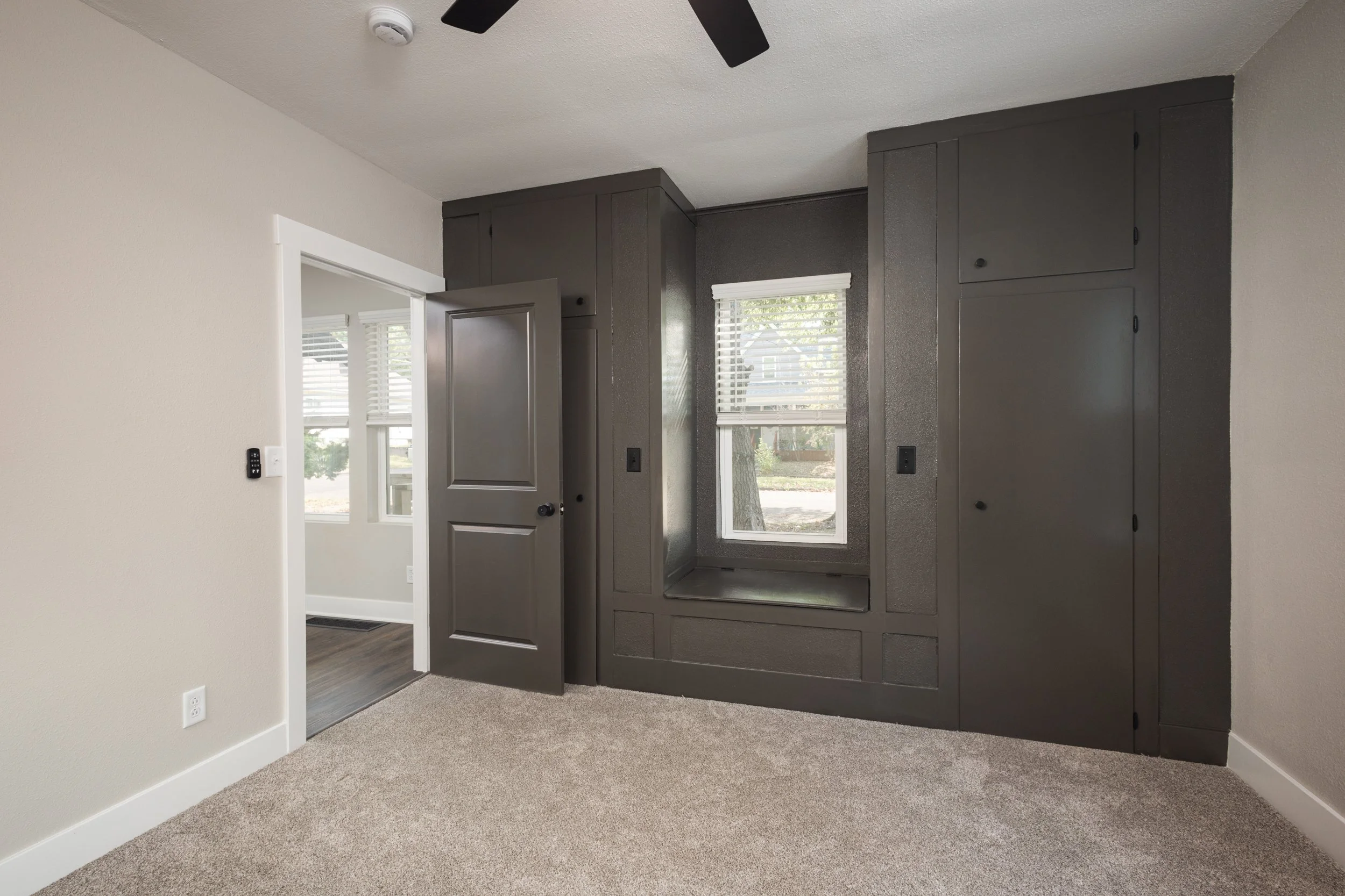 A room with beige carpet, beige walls, and a dark gray built-in wall unit with cabinets, a nook with a window, and a door. There is a ceiling fan with black blades and a smoke detector. An adjoing room with large windows and white blinds can be seen through an open doorway.