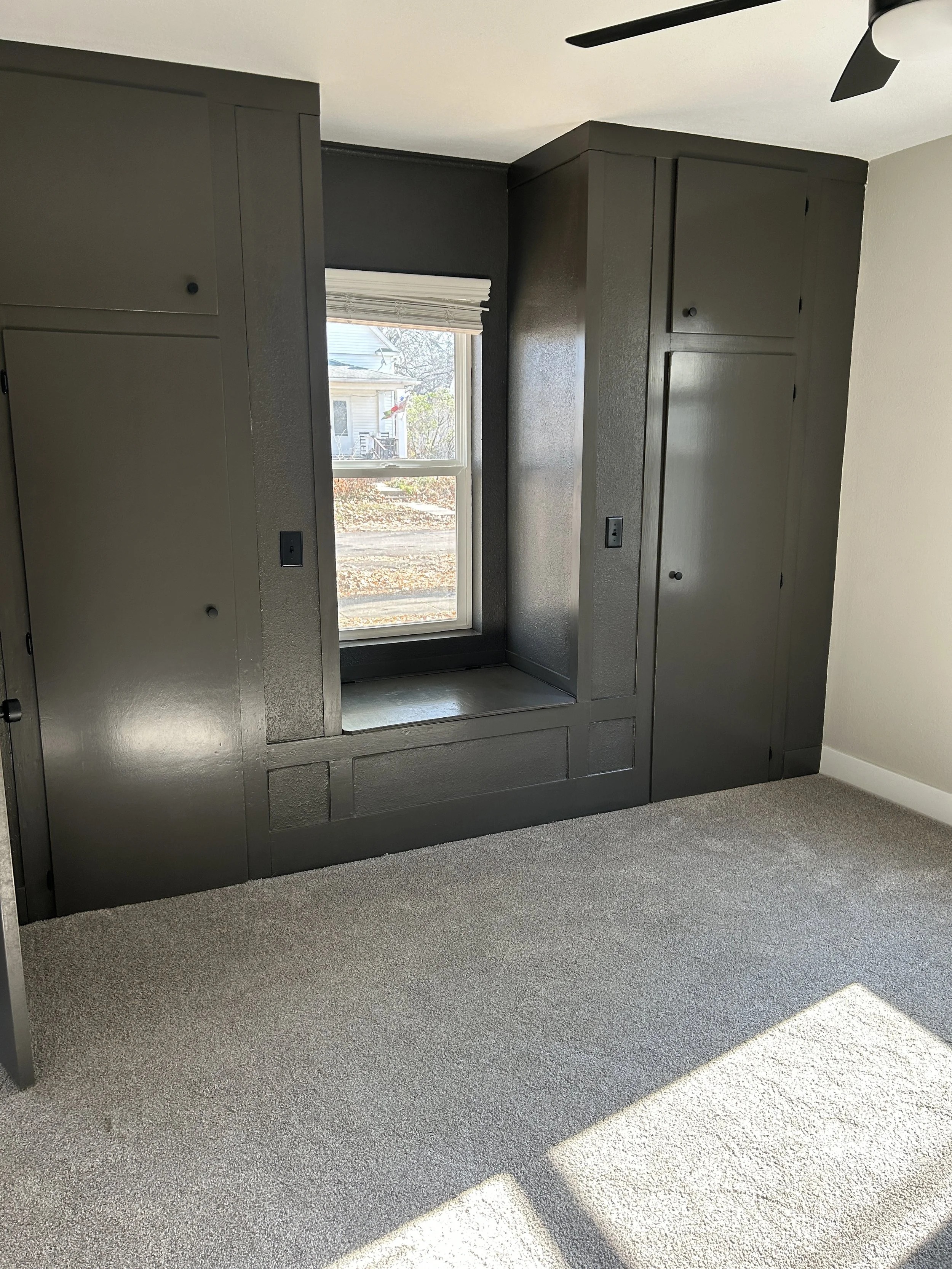 Interior of a room with dark gray built-in cabinets and a window with a view of a neighborhood outside.