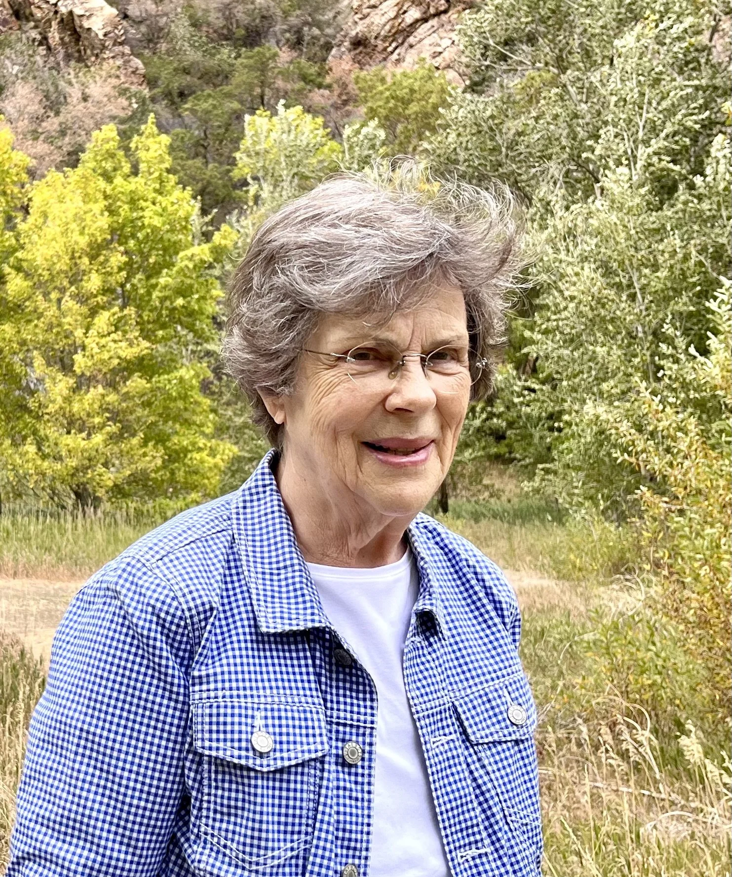 An elderly woman with short gray hair and glasses, smiling outdoors in a garden with trees and rocky hills in the background.