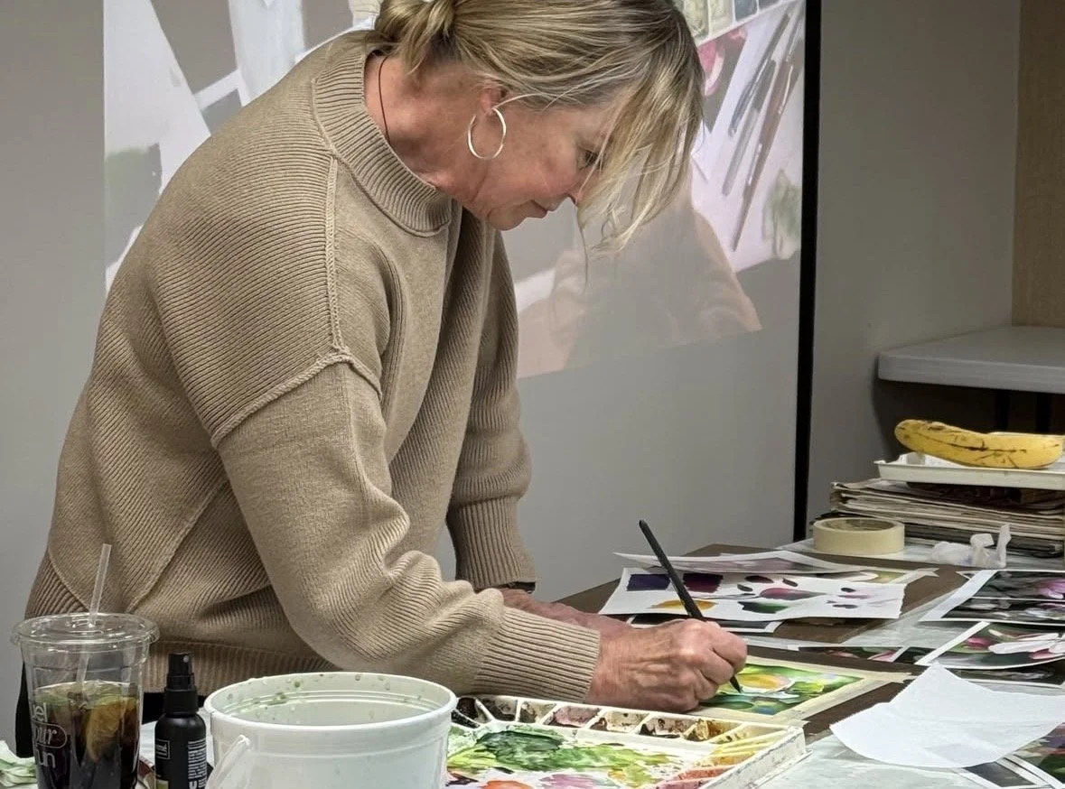 A woman with blonde hair in a beige sweater is painting with watercolors on paper, surrounded by various art supplies, on a table near a stack of papers and a banana.