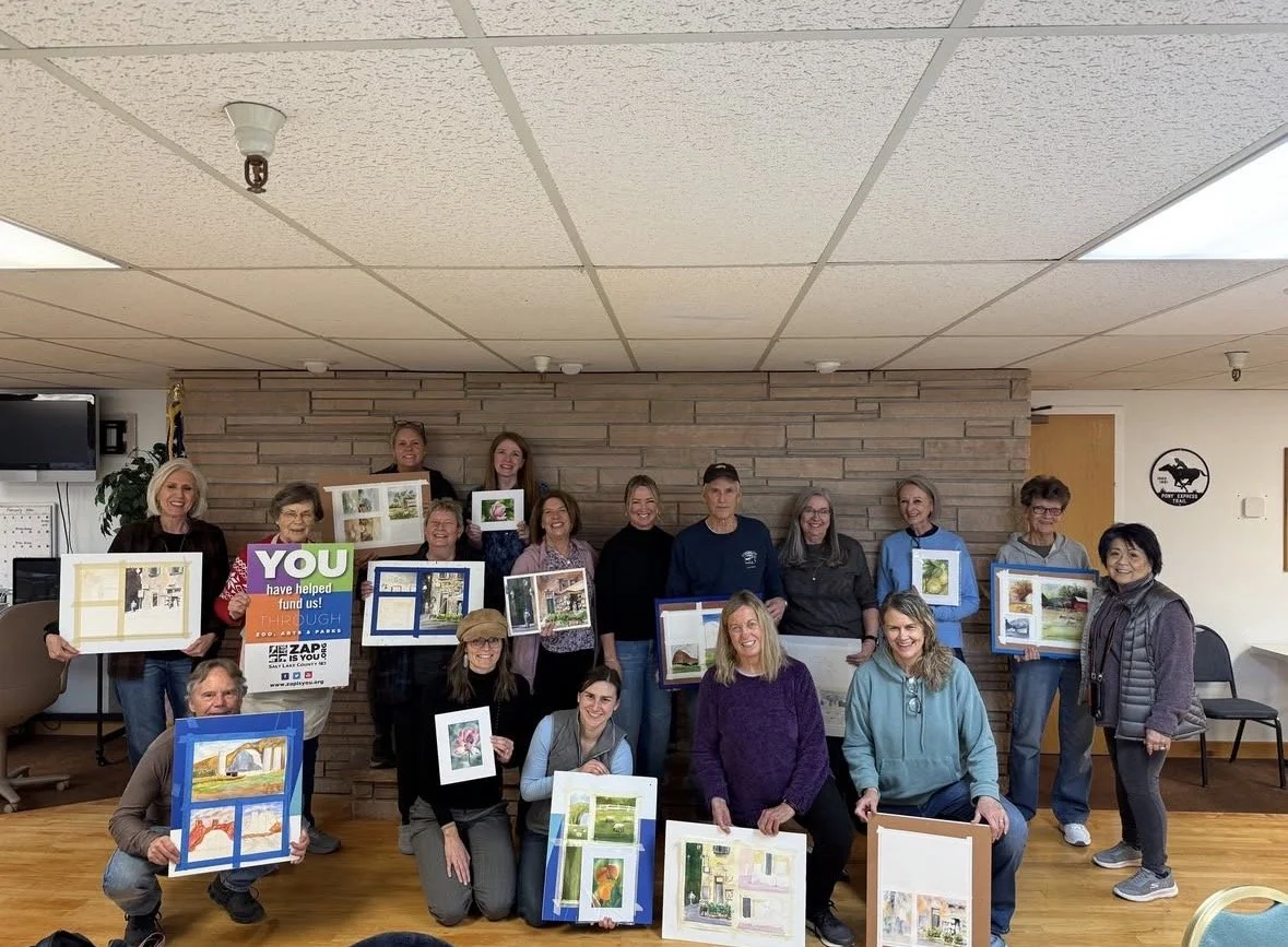 Group of people holding framed art projects and photos, standing and kneeling in a room with wood floor and brick wall.
