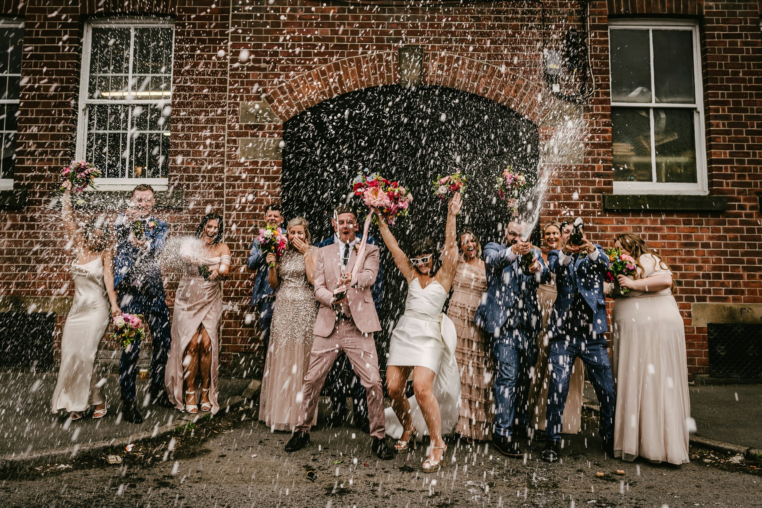 Hochzeitspaar und Gäste, die bei einer Feier vor einer Backsteinmauer mit Konfetti und Blumen feiern.