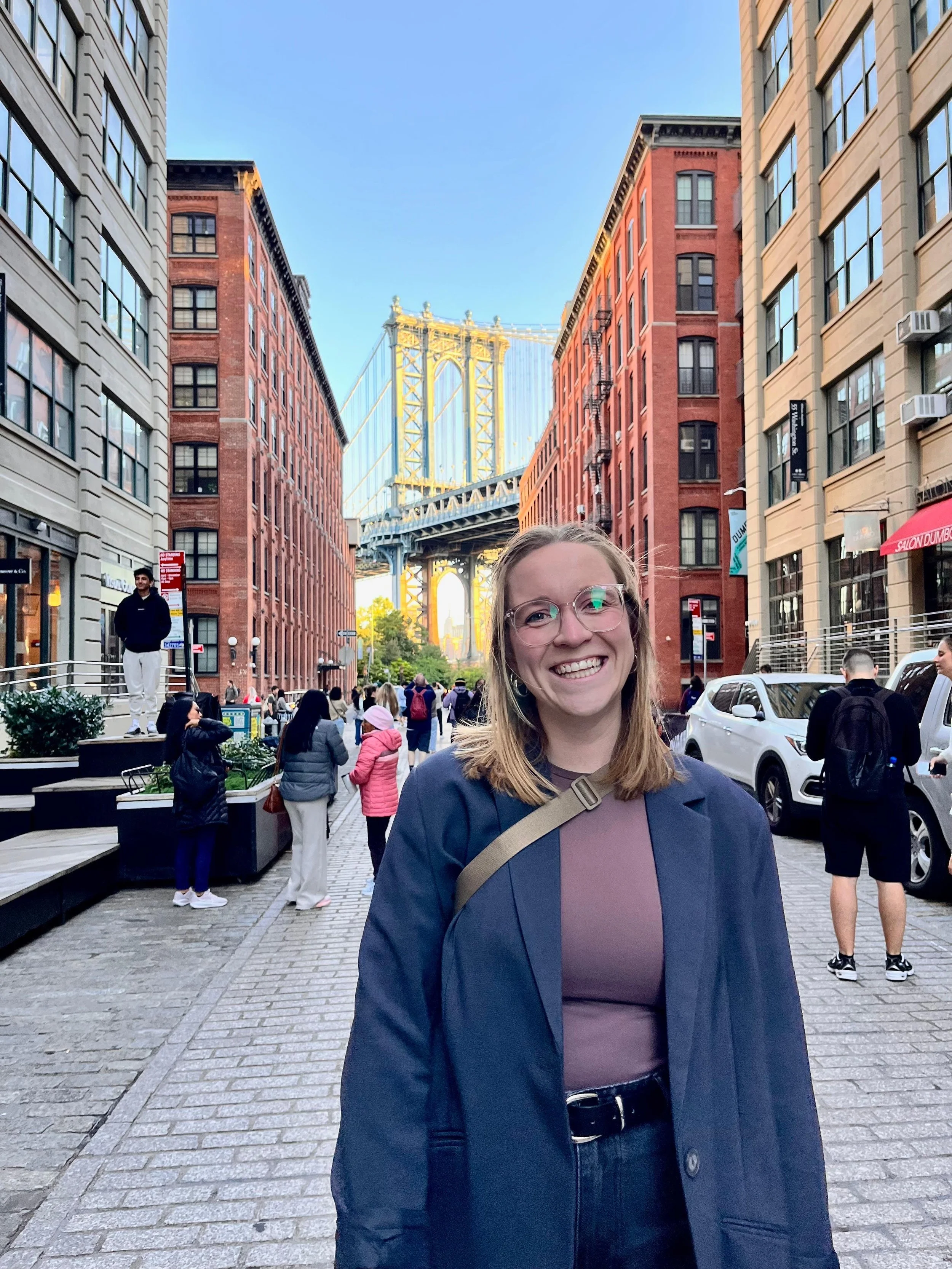 Eine lachende junge Frau mit Brille steht in einer belebten Stadtstraße mit Hochhäusern und Blick auf die Brooklyn Bridge im Hintergrund.
