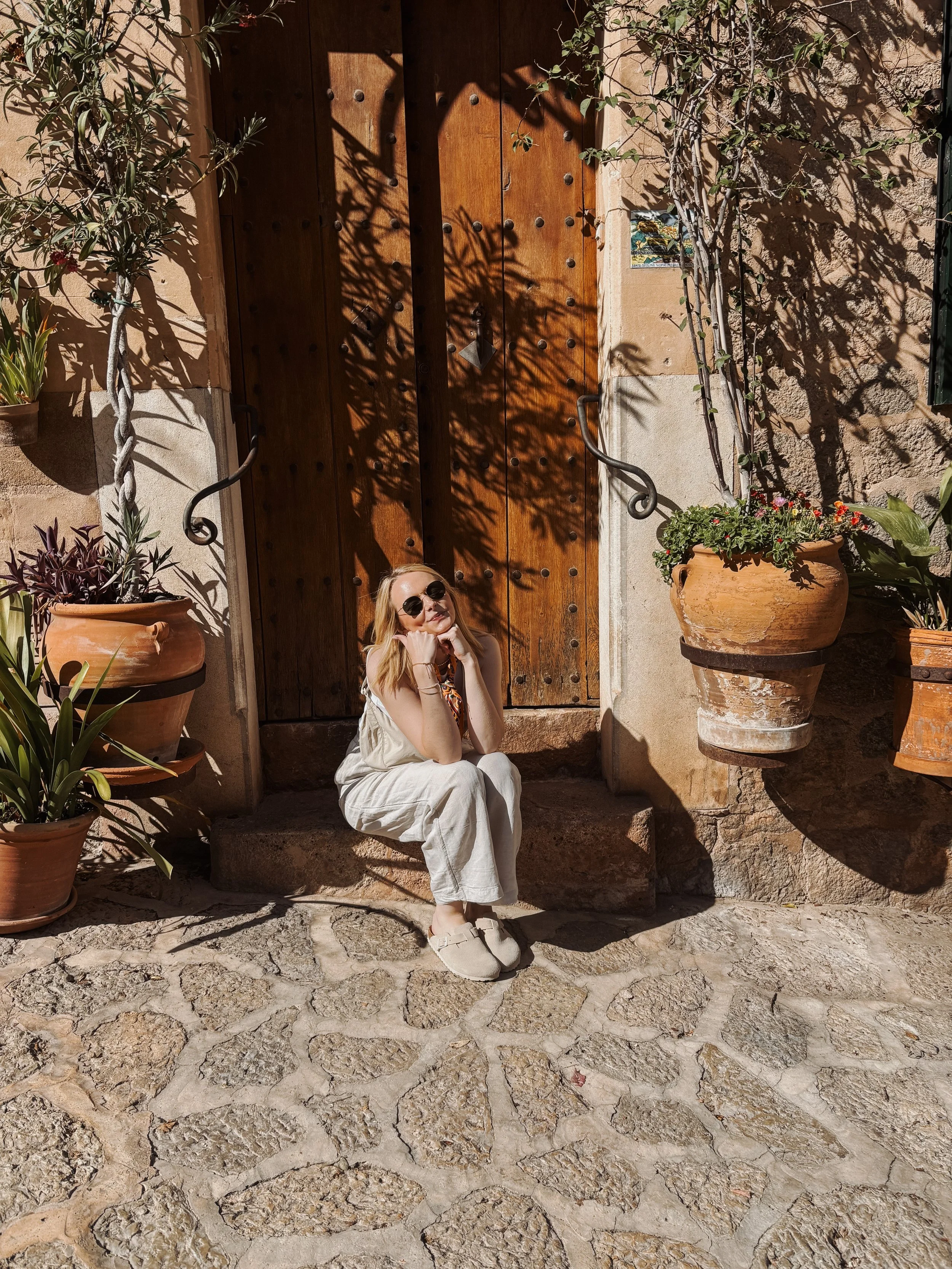 Eine junge Frau mit Sonnenbrille sitzt auf einer Steinstufe vor einem Holzportal, umgeben von Pflanzen in Terrakottatöpfen, bei Sonnenlicht im Freien.