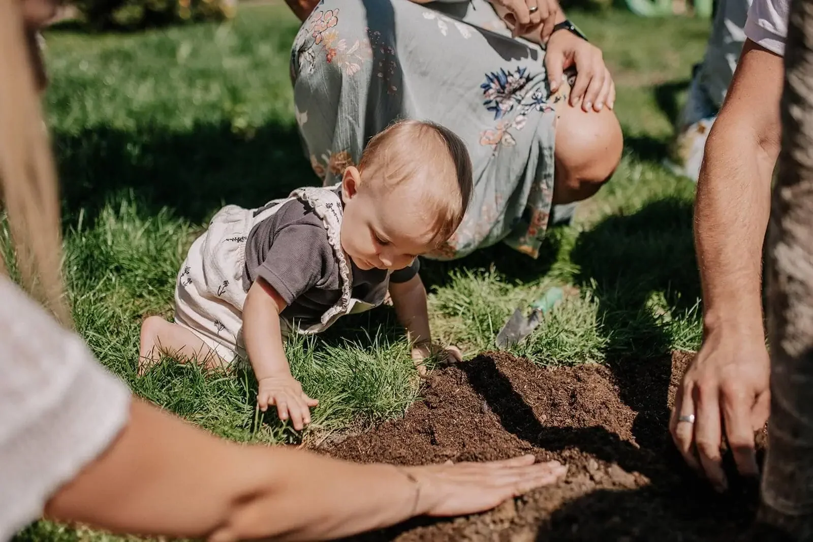 Ein Baby beim Pflanzen in einem Garten, umgeben von Erwachsenen, die beim Gärtnern helfen.