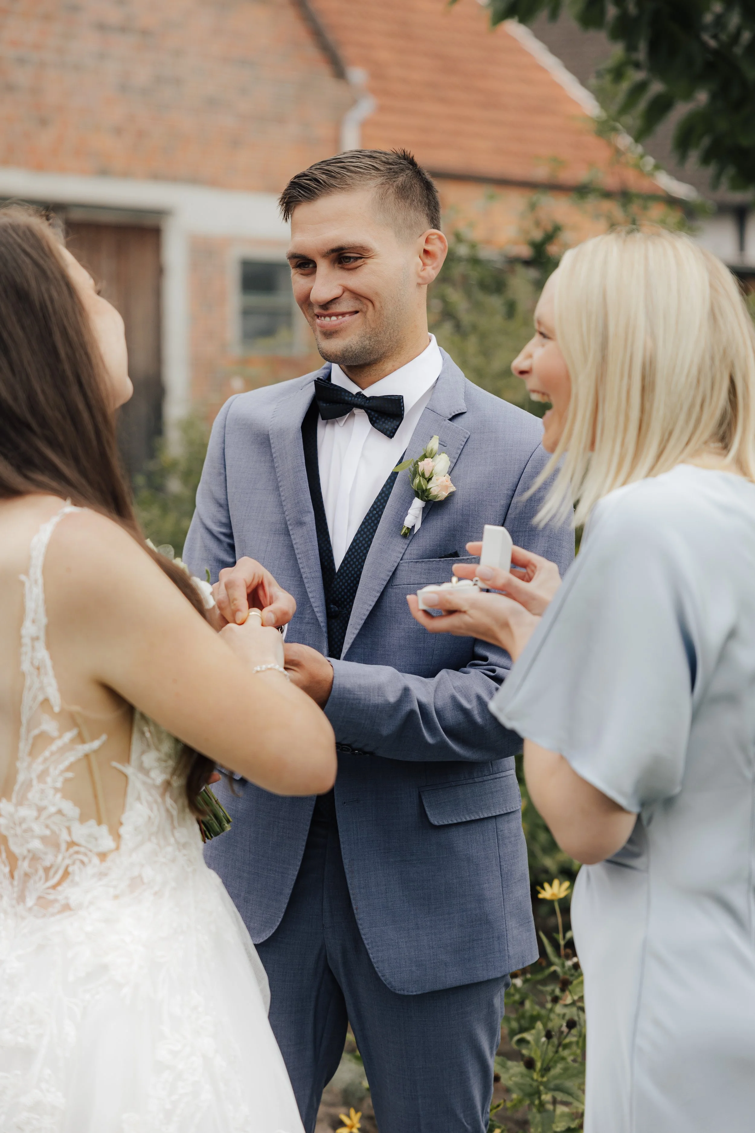 Ein Mann in einem grauen Anzug mit Fliege und eine Frau in einem weißen Kleid bei einer Hochzeitszeremonie, während eine andere Frau eine Ringdose hält und lächelt.