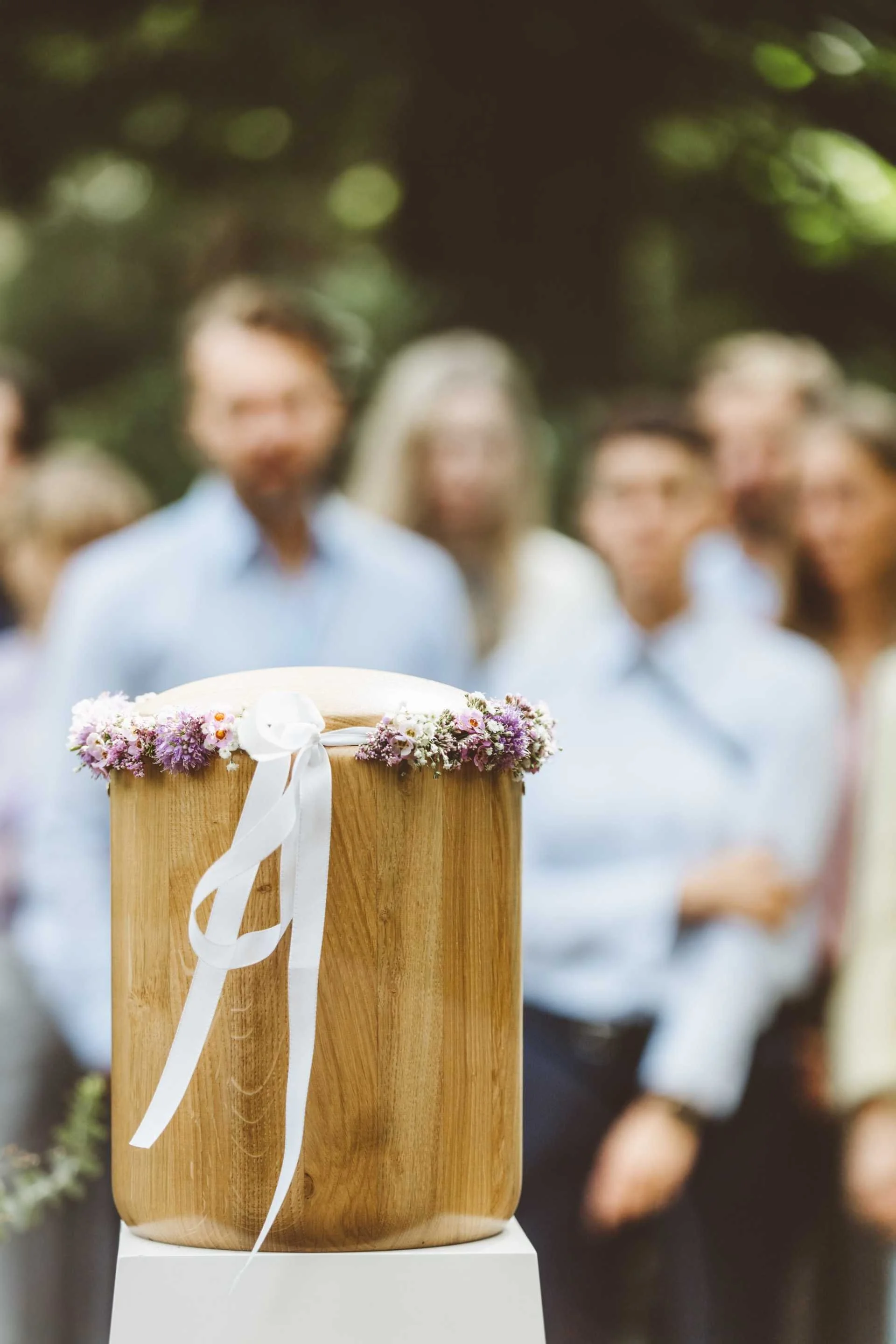 Eine traditionelle Taufschale aus Holz mit Blumenkranz auf einem Podest, im Hintergrund eine versammelte Gruppe Menschen bei einer Zeremonie im Freien.