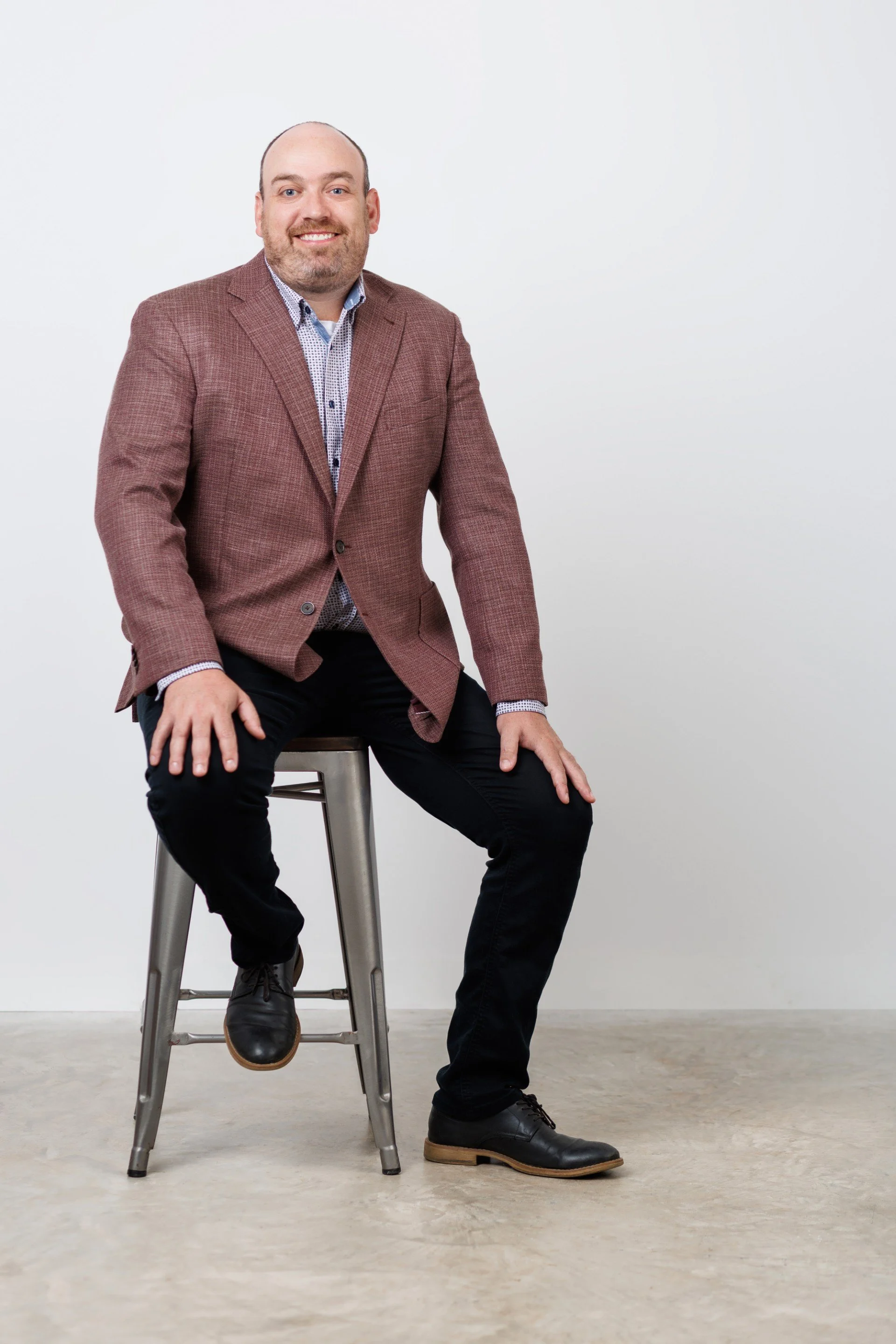A man with a beard and bald head wearing a brown blazer, light patterned shirt, black pants, and black shoes sitting on a silver chair against a plain white background.