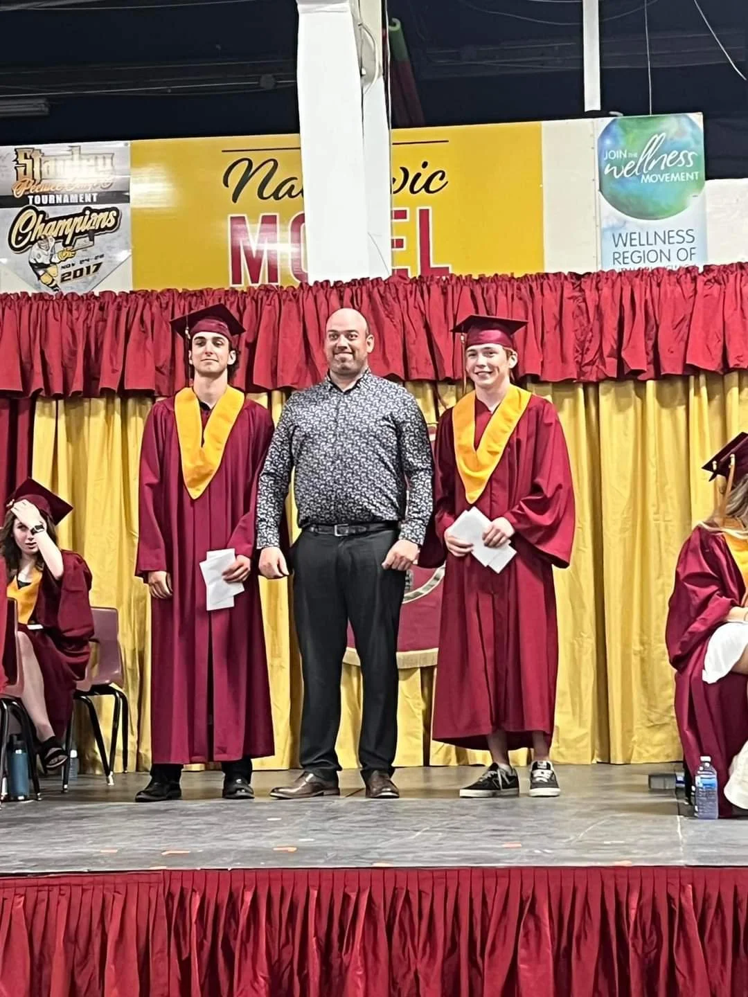 Group of students and a man standing on a stage during a graduation ceremony. The students are in maroon caps and gowns with yellow sashes. The background has red and yellow curtains and signs, including one that says "National Model" and another promoting Wellness Movement.