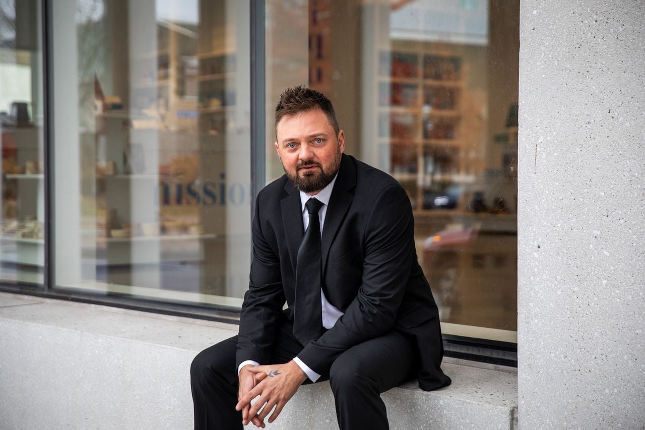 Man in a black suit and tie sitting on a concrete ledge outside of a storefront with large windows.