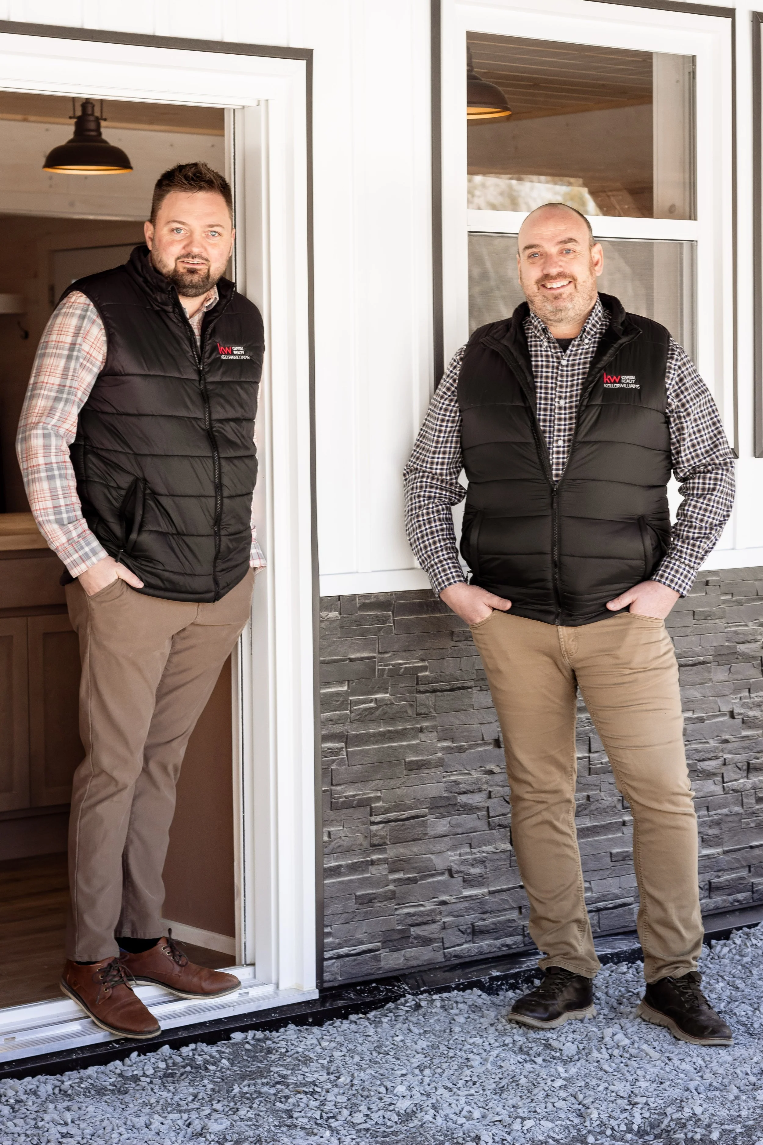 Two men standing outside a building, both wearing black vests over plaid shirts and khaki pants. One is standing in a doorway, and the other is near a window, with a textured stone wall behind them.
