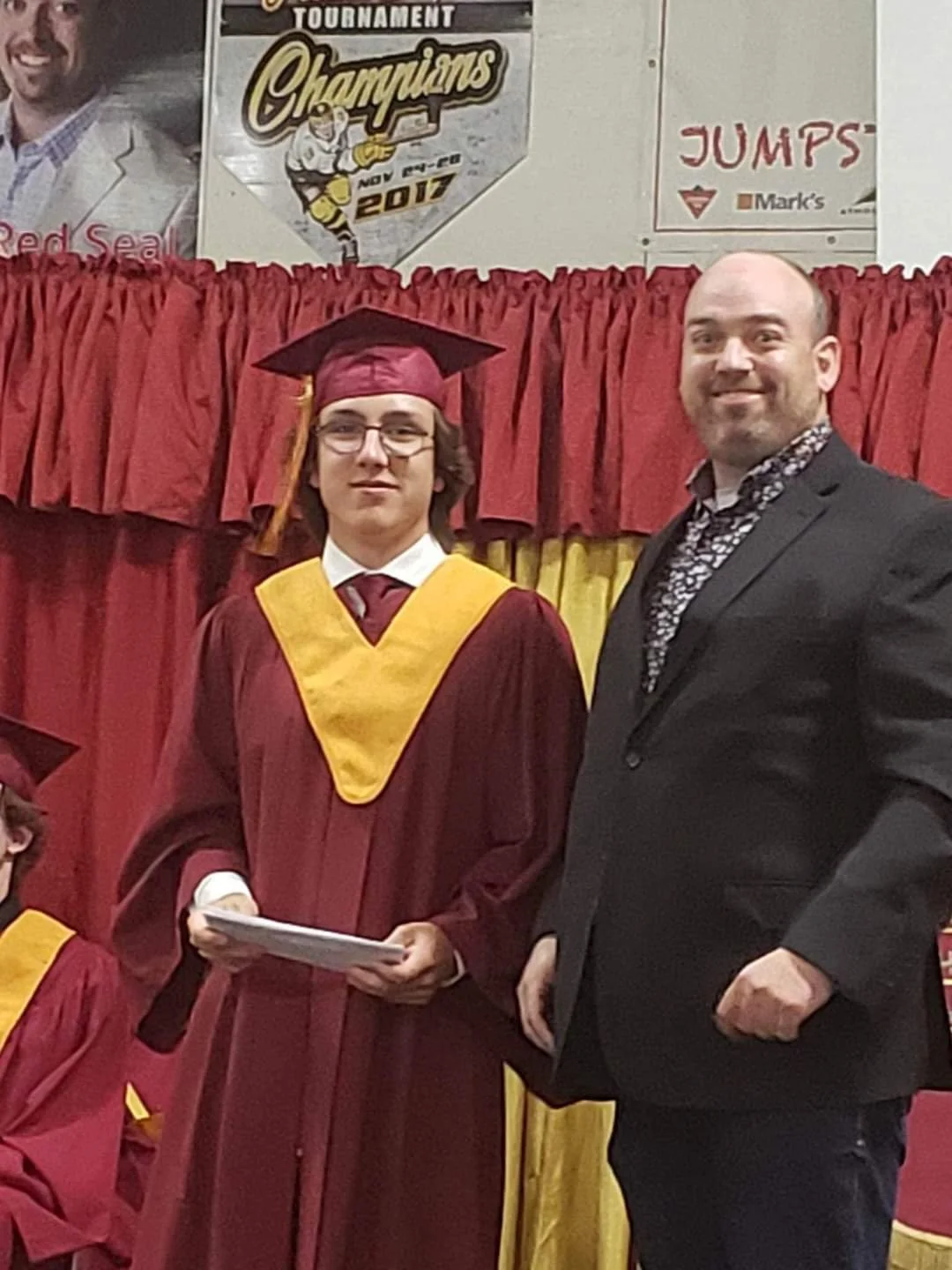 A young man in a maroon graduation gown and cap with a yellow stole standing next to an older man in a black suit at a graduation ceremony. There are red curtains behind them and a banner above them celebrating a hockey tournament champions from 2017.