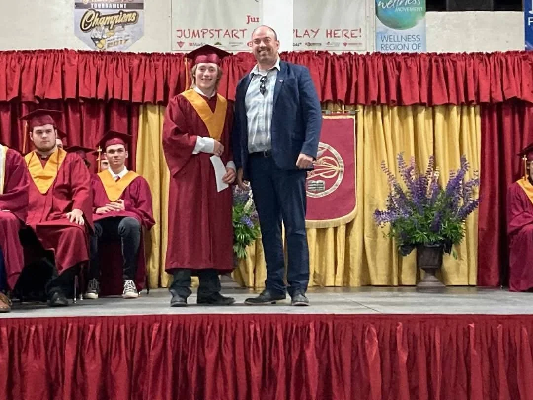 A graduation ceremony on stage with students in maroon caps and gowns with gold accents, and an adult man in a suit. One student is standing and receiving a diploma, smiling, while others sit or stand nearby.