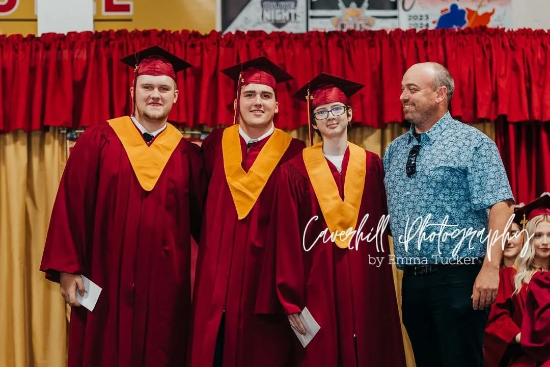Three graduates in red caps and gowns with yellow stoles, standing with a man at a graduation ceremony. Red curtain backdrop.