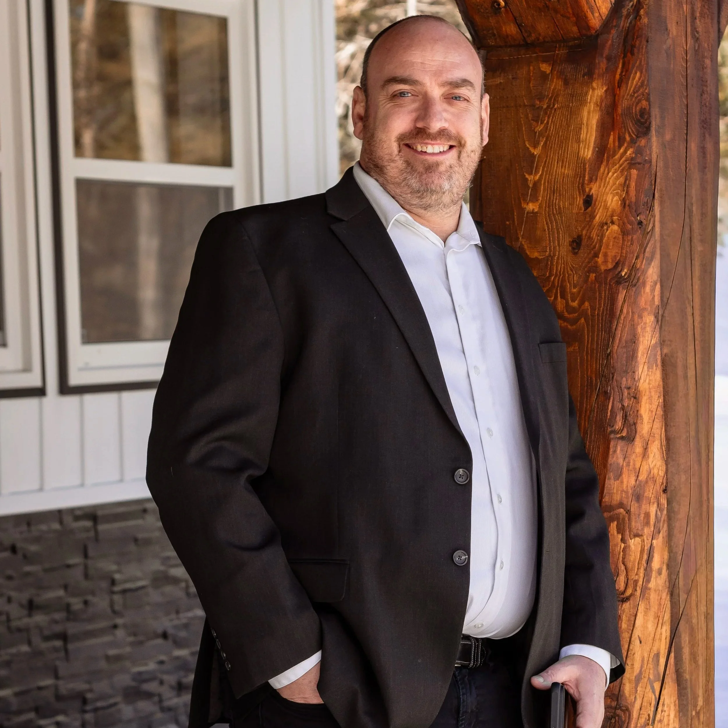 A man in a black suit and white shirt standing outside the house, leaning against a wooden post, smiling.