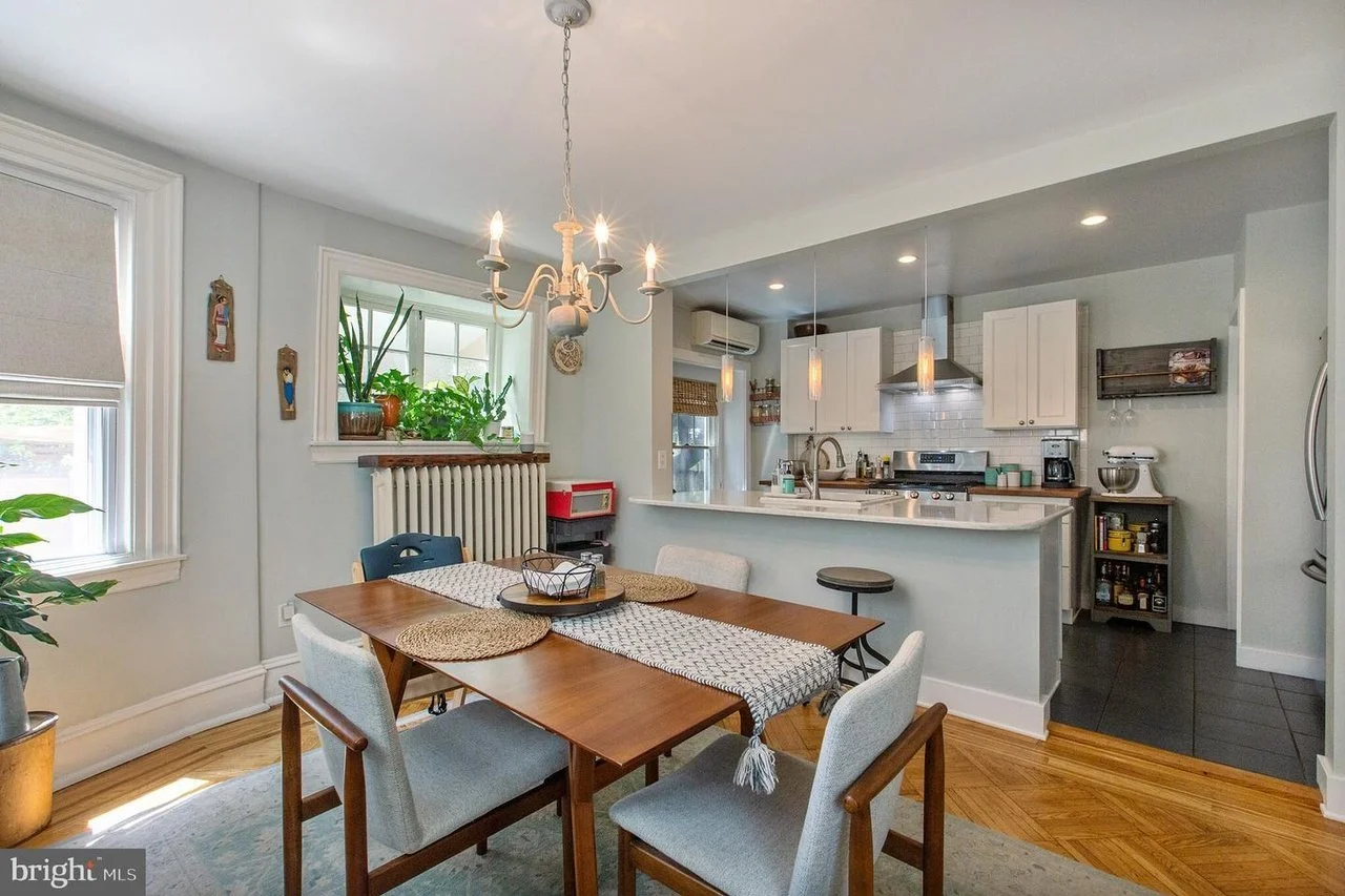 A dining area with a wooden table, six chairs, and a chandelier, adjacent to a kitchen with white cabinets, stainless steel appliances, and a breakfast bar.