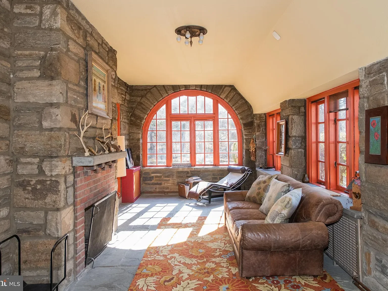 Living room with large arched window, stone walls, and red window frames, featuring a brown leather sofa, decorative pillows, a colorful area rug, a fireplace with antlers on the mantle, and a chaise lounge near the window.