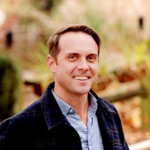 A man outdoors with a blurred autumnal background, smiling, wearing a light blue button-up shirt and a dark jacket.