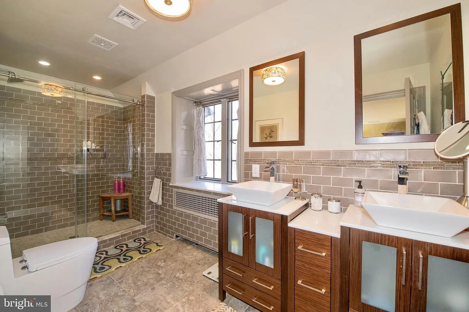 Modern bathroom with double vanity sinks, two mirrors with wooden frames, a window, and a walk-in shower with tiled walls and glass doors.
