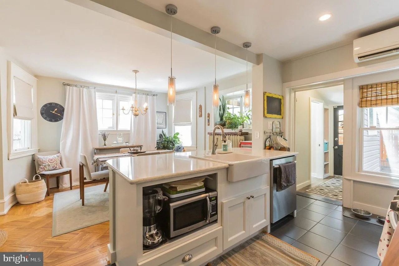 Bright open-concept kitchen and dining area with white cabinets, pendant lighting, and a marble kitchen island. The dining area has a table, chairs, and a chandelier. Windows with curtains and plants adorn the space.