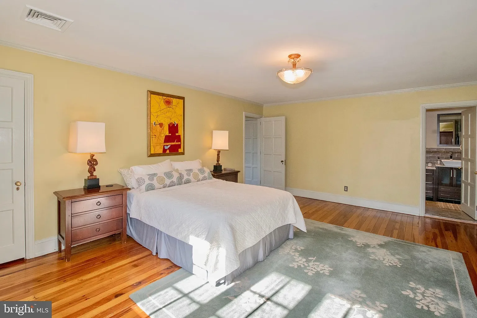A bedroom with a bed, two bedside tables with lamps, yellow walls, and a hardwood floor. There's a painting above the bed and a doorway leading to a bathroom.