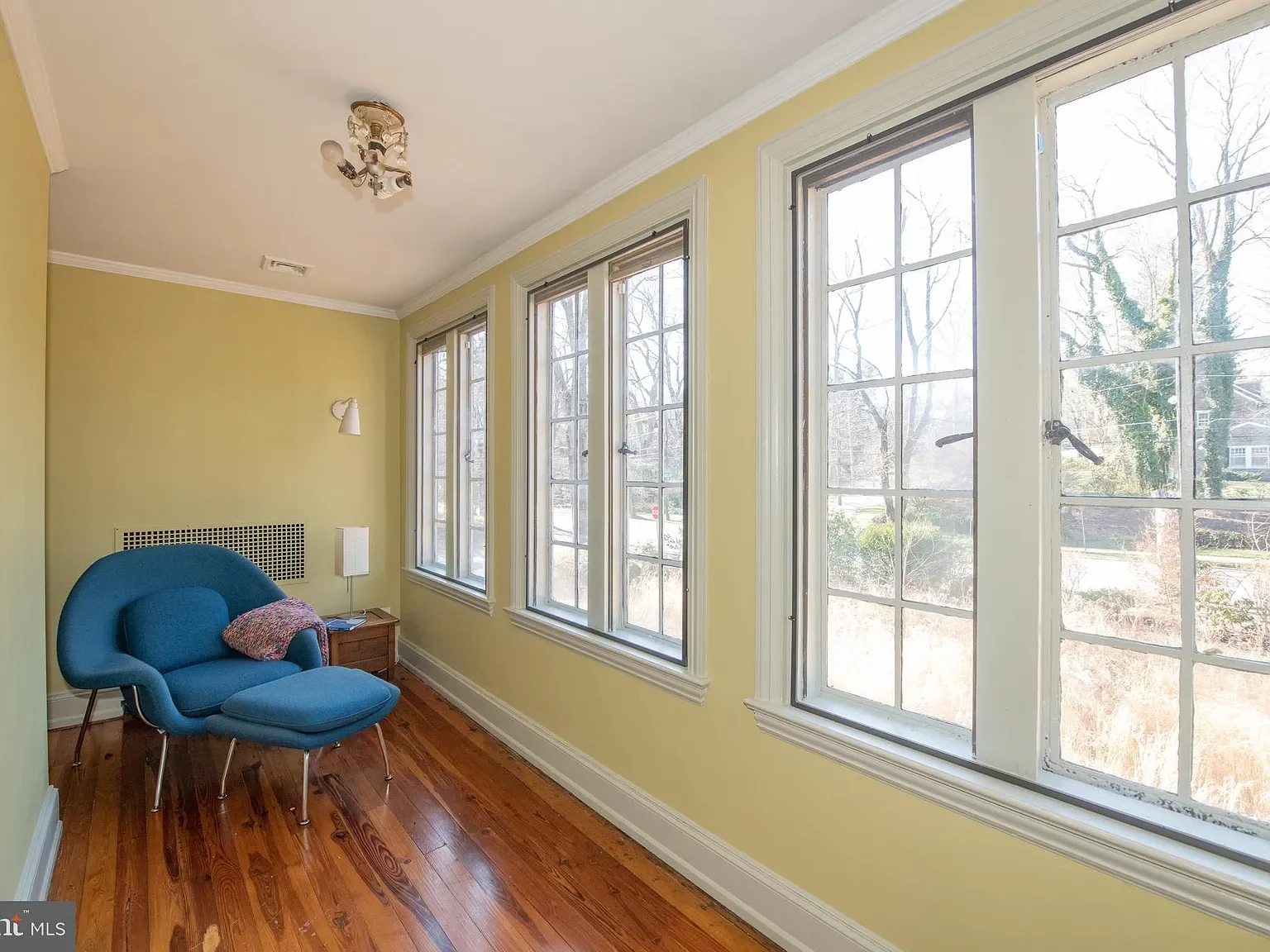 A cozy corner in a sunlit room with yellow walls, featuring a blue armchair with an ottoman, a side table, and large windows showing trees outside.