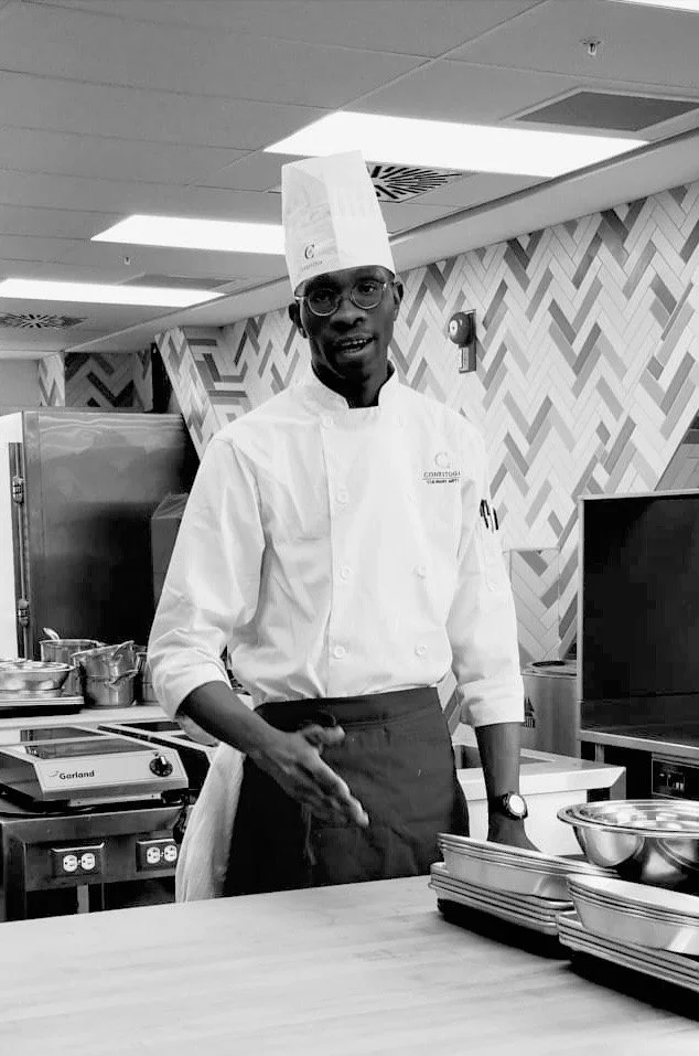 A male chef in a white chef's coat and tall chef's hat standing in a kitchen with stacked trays and cooking equipment, smiling and gesturing with his hand.