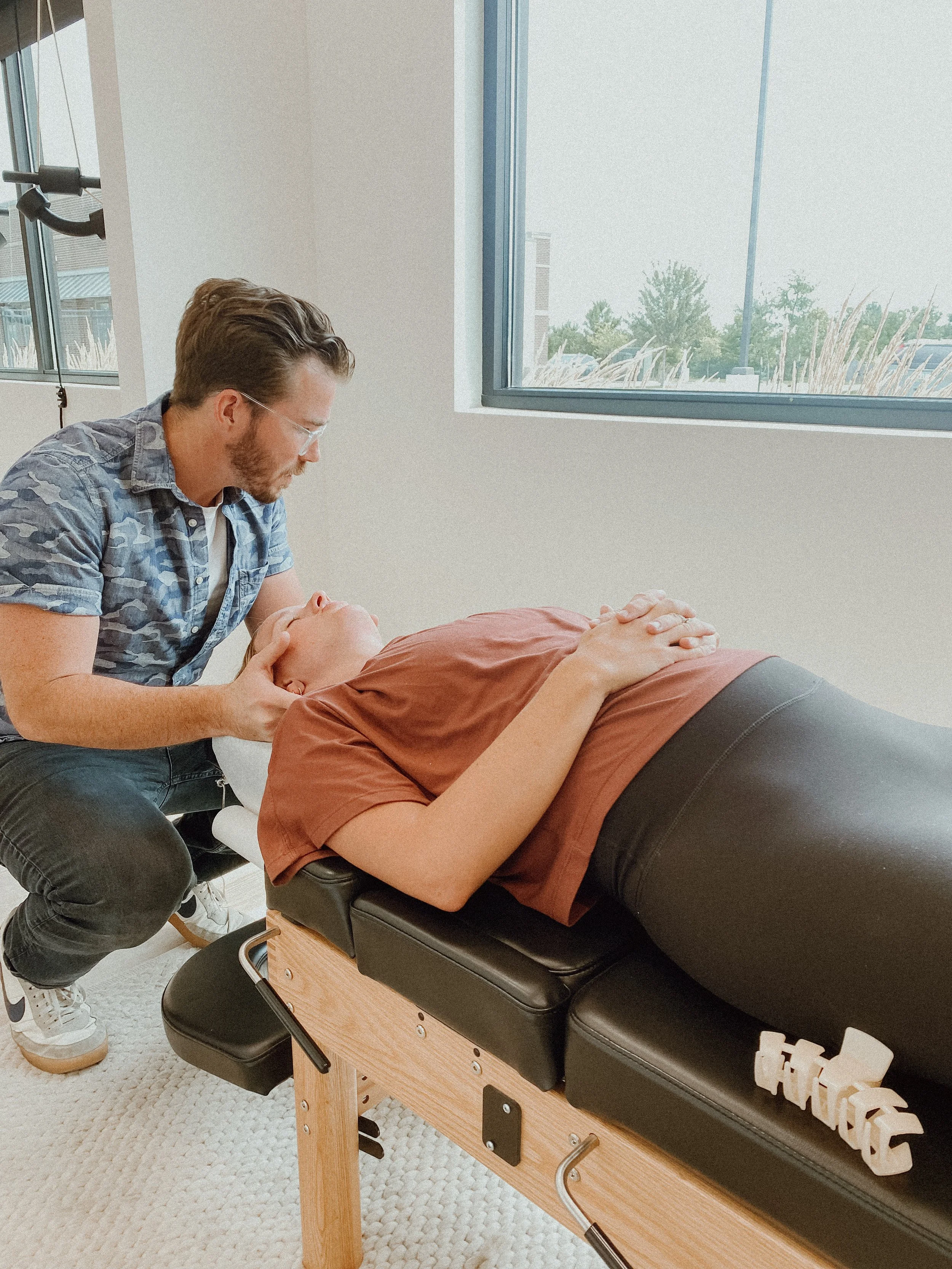 A chiropractor adjusting a patient lying face-up on a chiropractic table in a well-lit room with large windows.