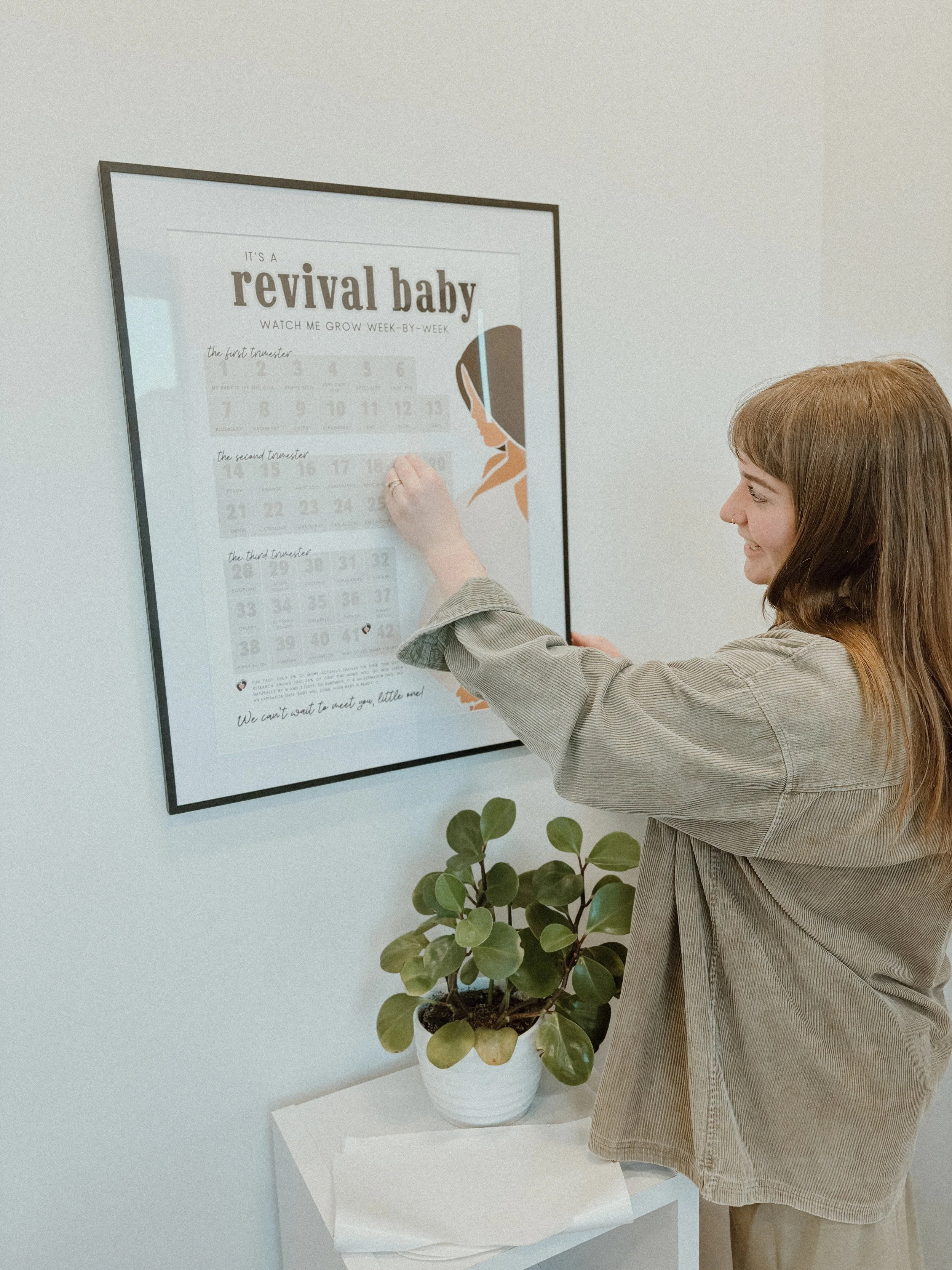 A woman with shoulder-length brown hair, smiling as she points to a calendar on the wall. The calendar is titled 'It's a revival baby' and features an illustration of a woman. A houseplant in a white pot sits on a white stand below the calendar.