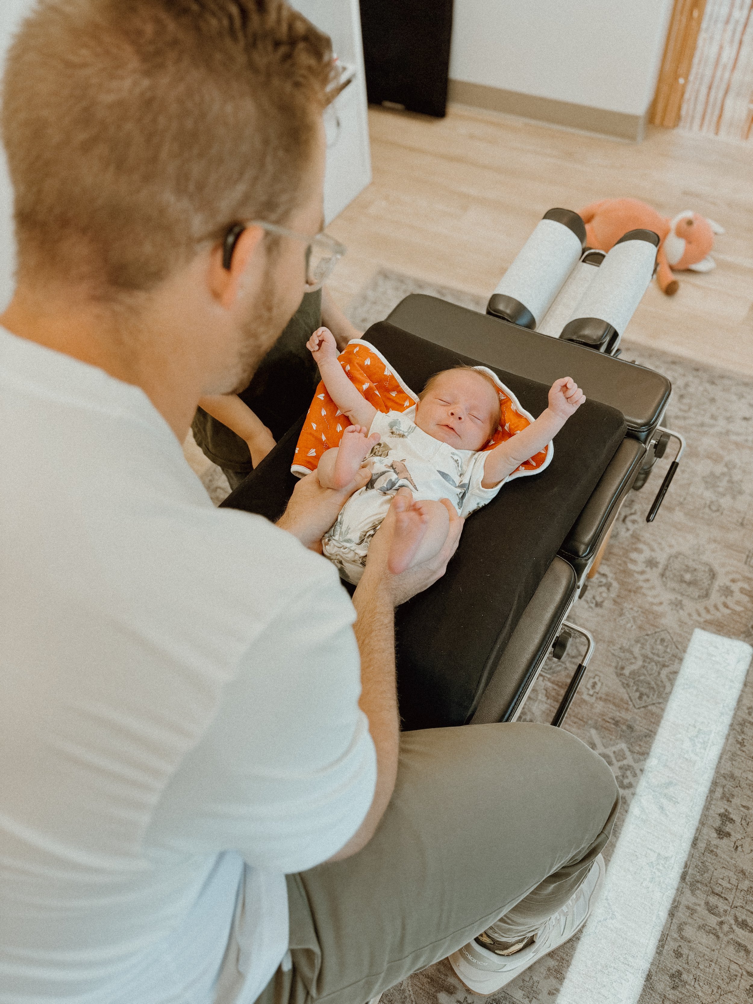 A man kneeling beside a baby lying on a changing table, looking at the baby with arms raised in a stretch, in a room with a wooden floor and plush toys in the background.